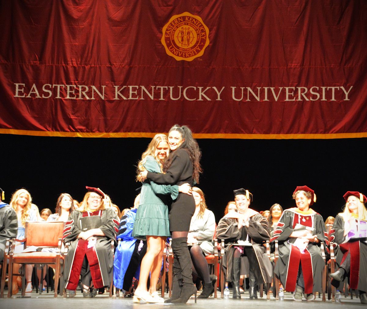 Two women hugging on stage at Eastern Kentucky University graduation. Banner with university logo in background.