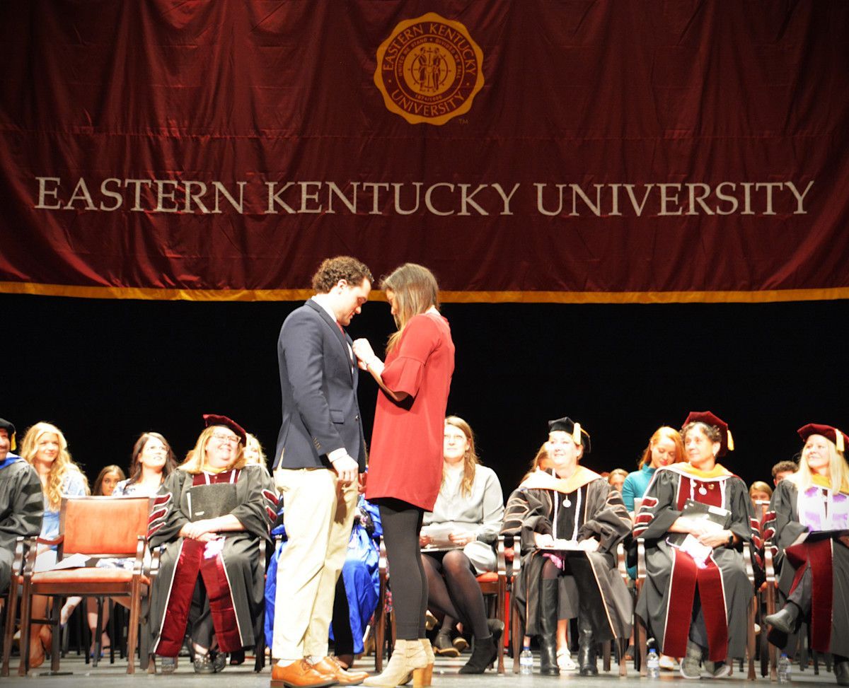 A person in a red dress pins a badge on a person in a suit on stage at Eastern Kentucky University graduation.