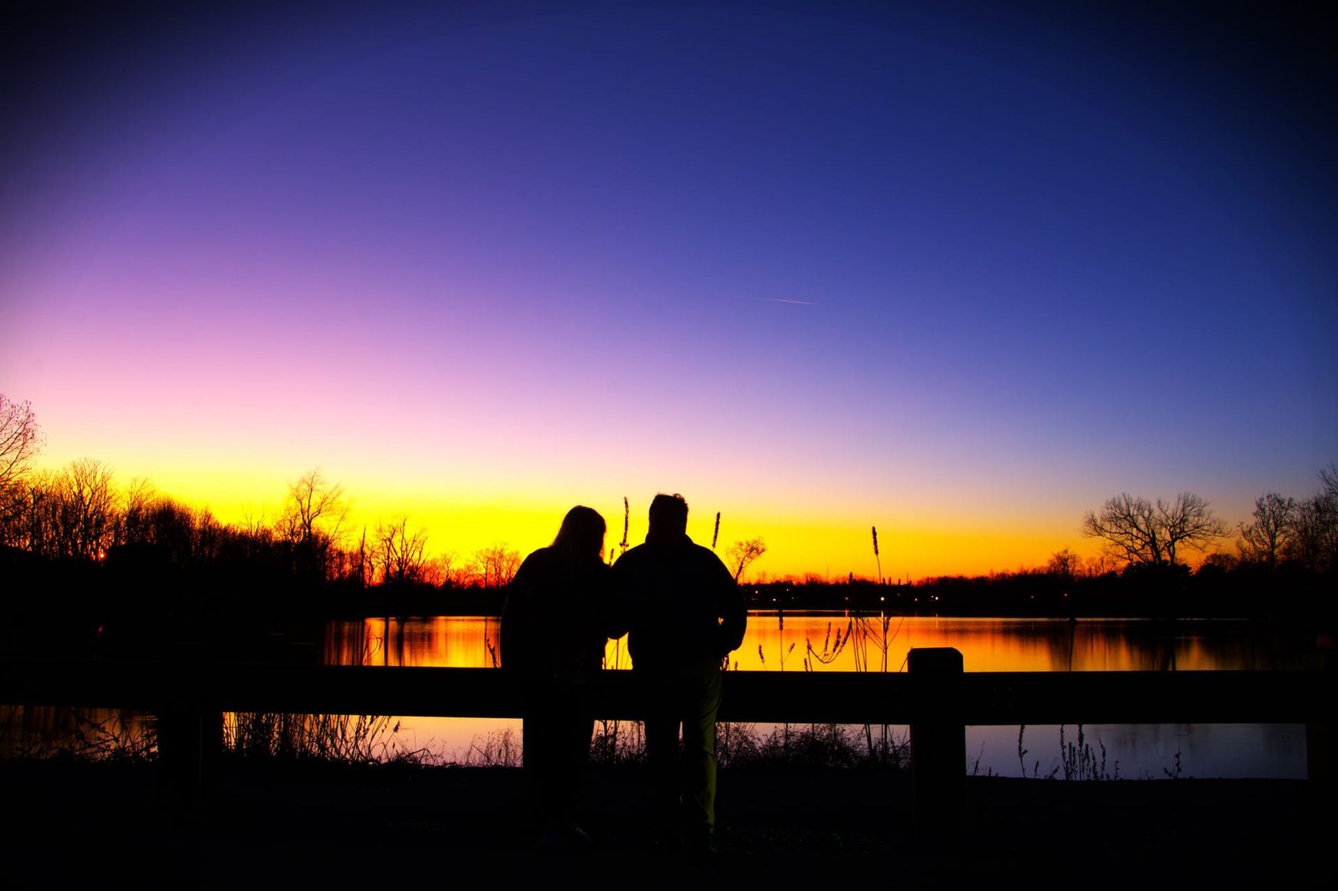 Two silhouetted people watch a colorful sunset over a lake.