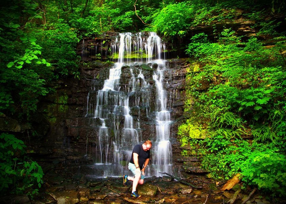Man kneels near tiered waterfall in lush green forest.