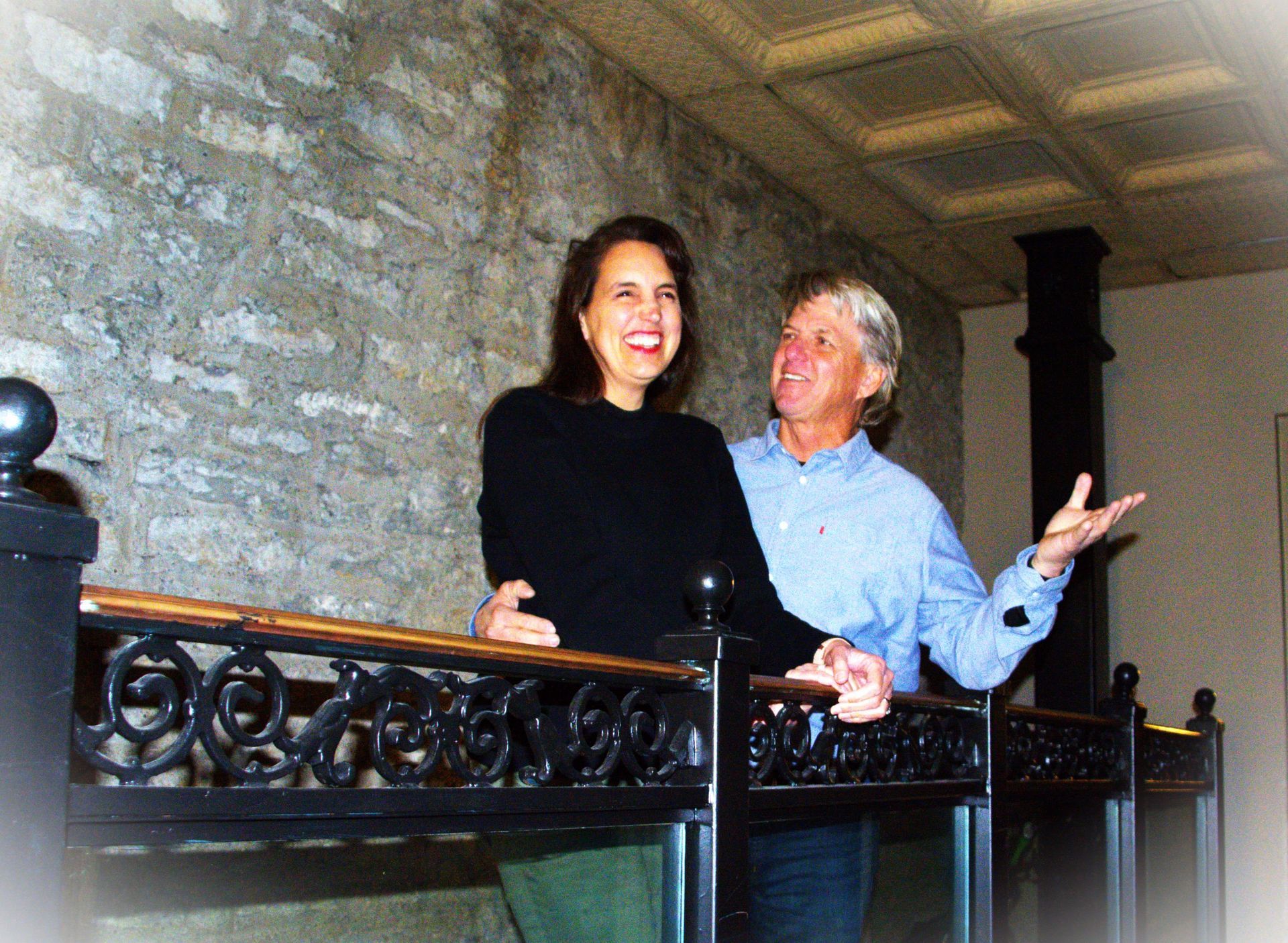 Woman and man on a balcony, smiling. The stone wall and decorative railing, with a white patterned ceiling.