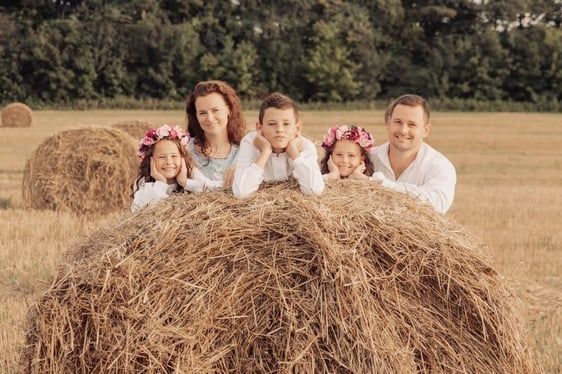 Family poses on a hay bale in a field. Smiling, wearing white shirts, flower crowns.