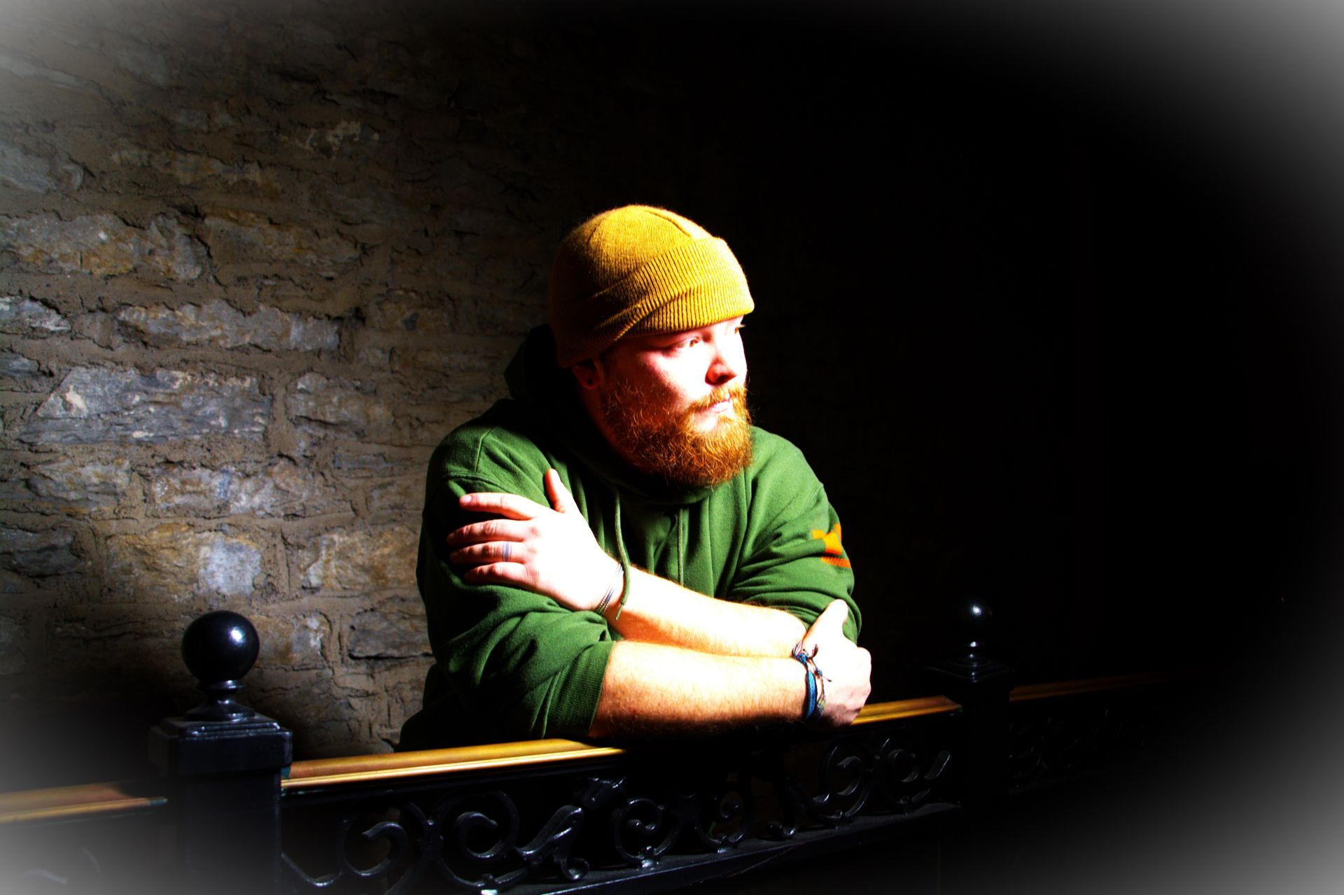 Man with a red beard and yellow hat leans on a dark railing, looking away; wearing green hoodie, against stone wall.