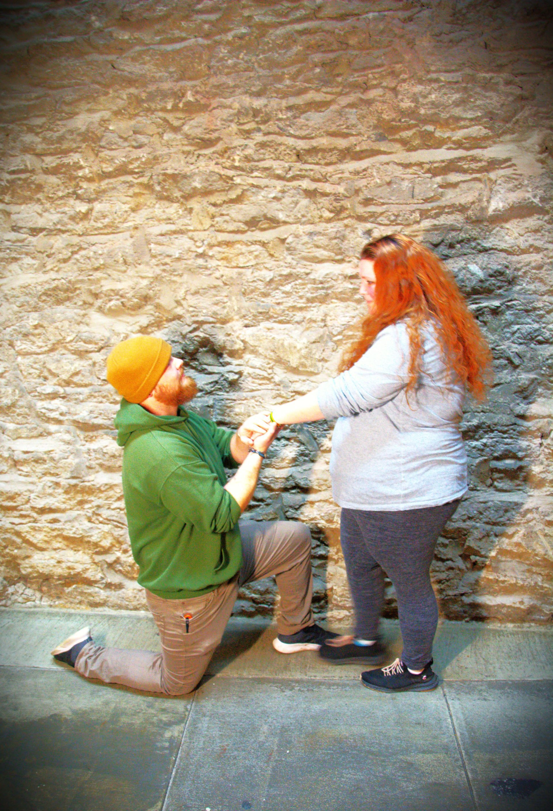 Man on one knee, holding woman's hands, proposing in front of a stone wall.
