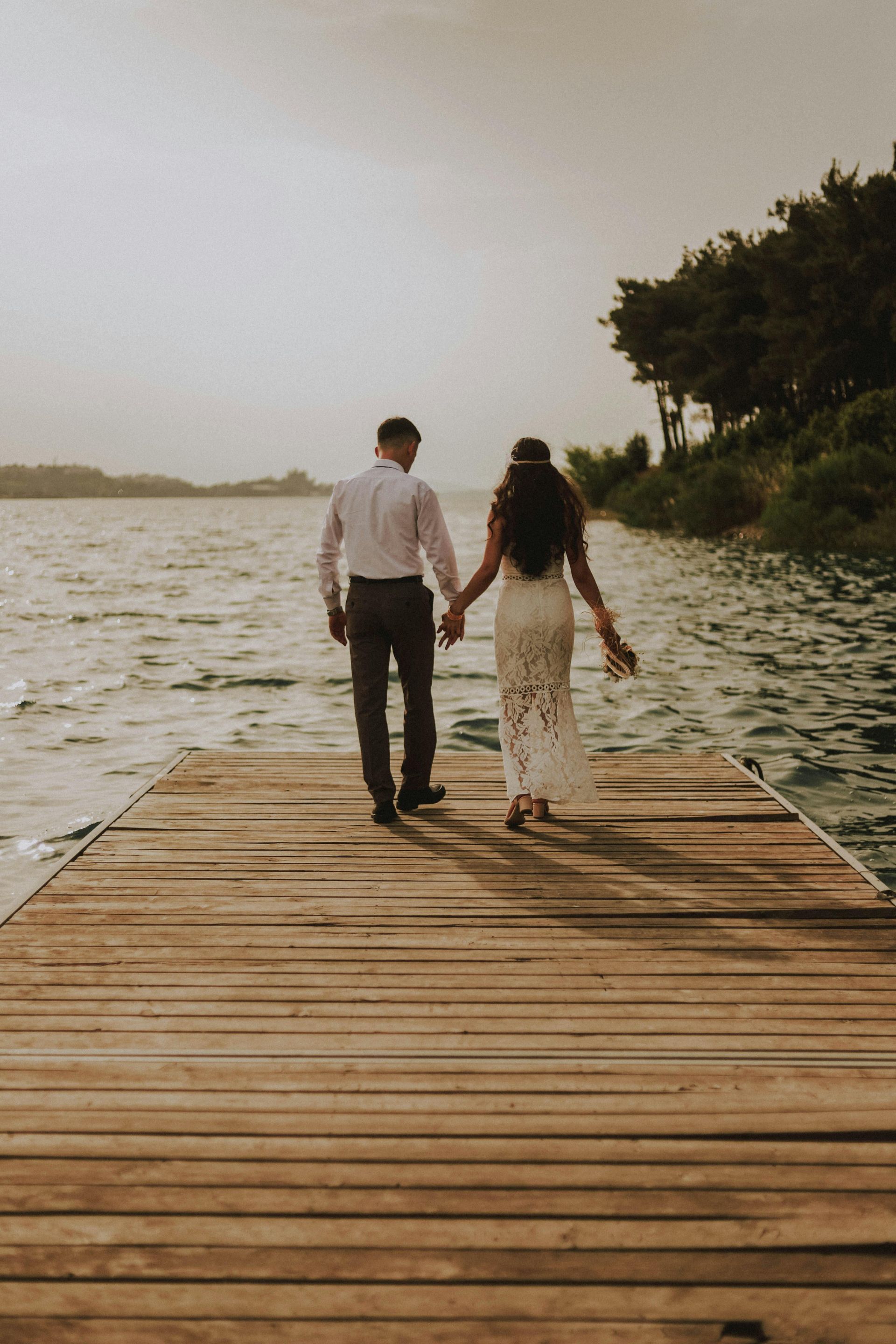 Couple holding hands, walking on a wooden pier towards a lake, with trees in the background.
