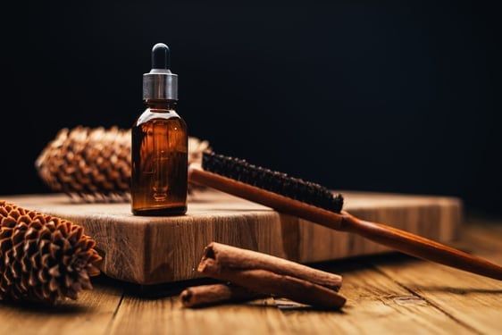 Amber bottle with dropper, brush, pine cones, and cinnamon sticks on a wooden surface.