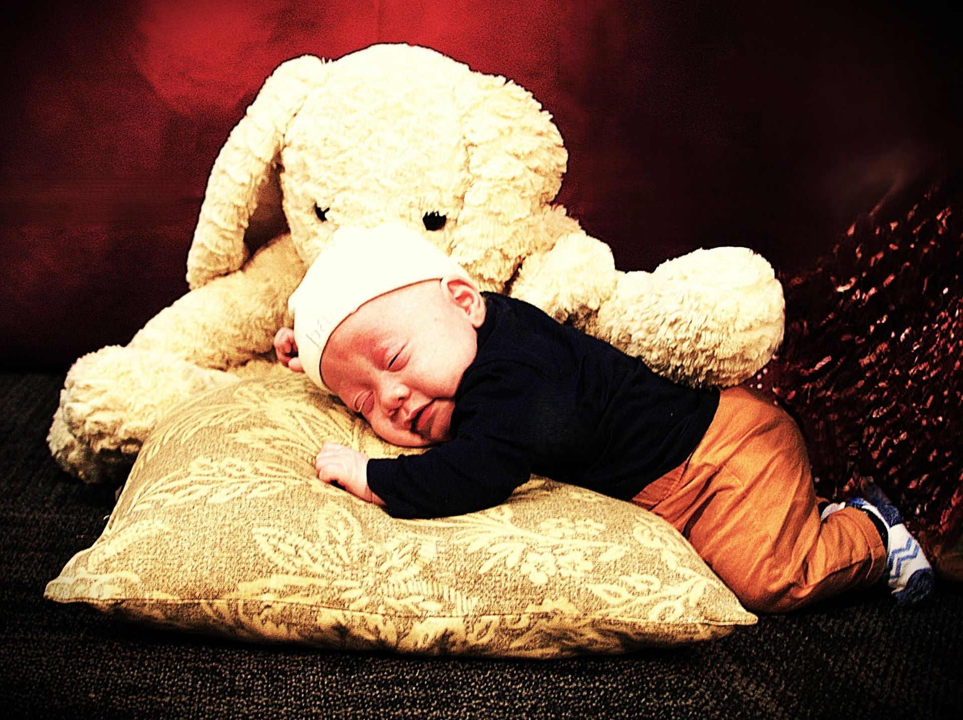 Sleeping baby in a white hat and orange pants rests on a pillow with a large stuffed rabbit.