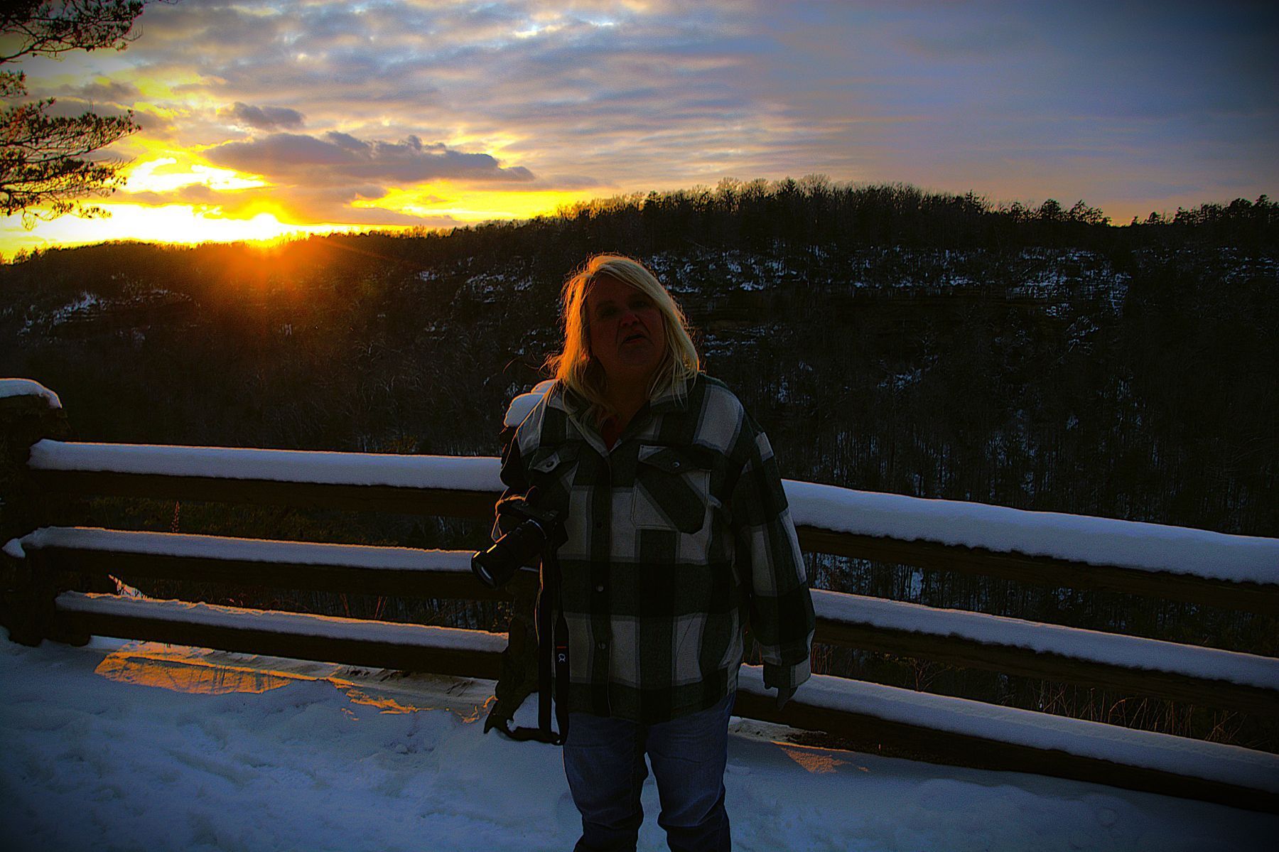Person standing in snow-covered area, looking at sunset over trees. Wearing plaid jacket, holding object.