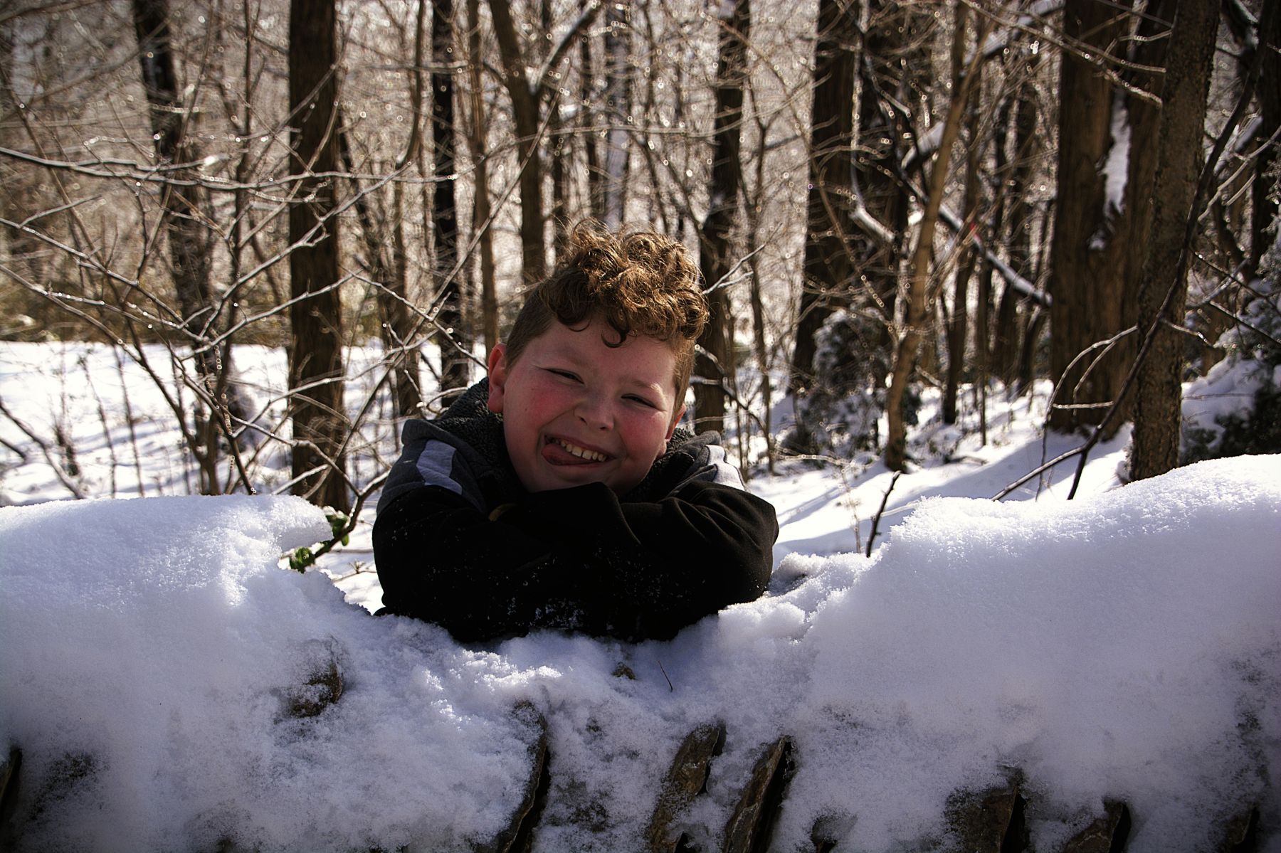 A person with curly hair smiles while leaning over a snow-covered wooden fence in a snowy forest.