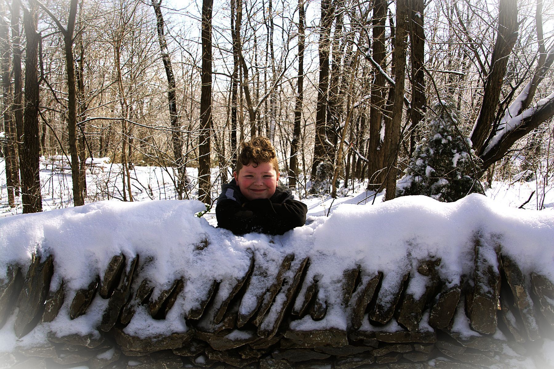 Child smiling behind snow-covered stone wall in a snowy forest.