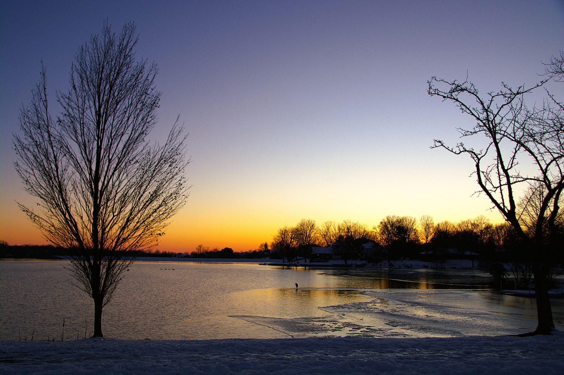 Sunset over a lake; bare trees frame the water. Orange and purple sky. Some ice and snow.