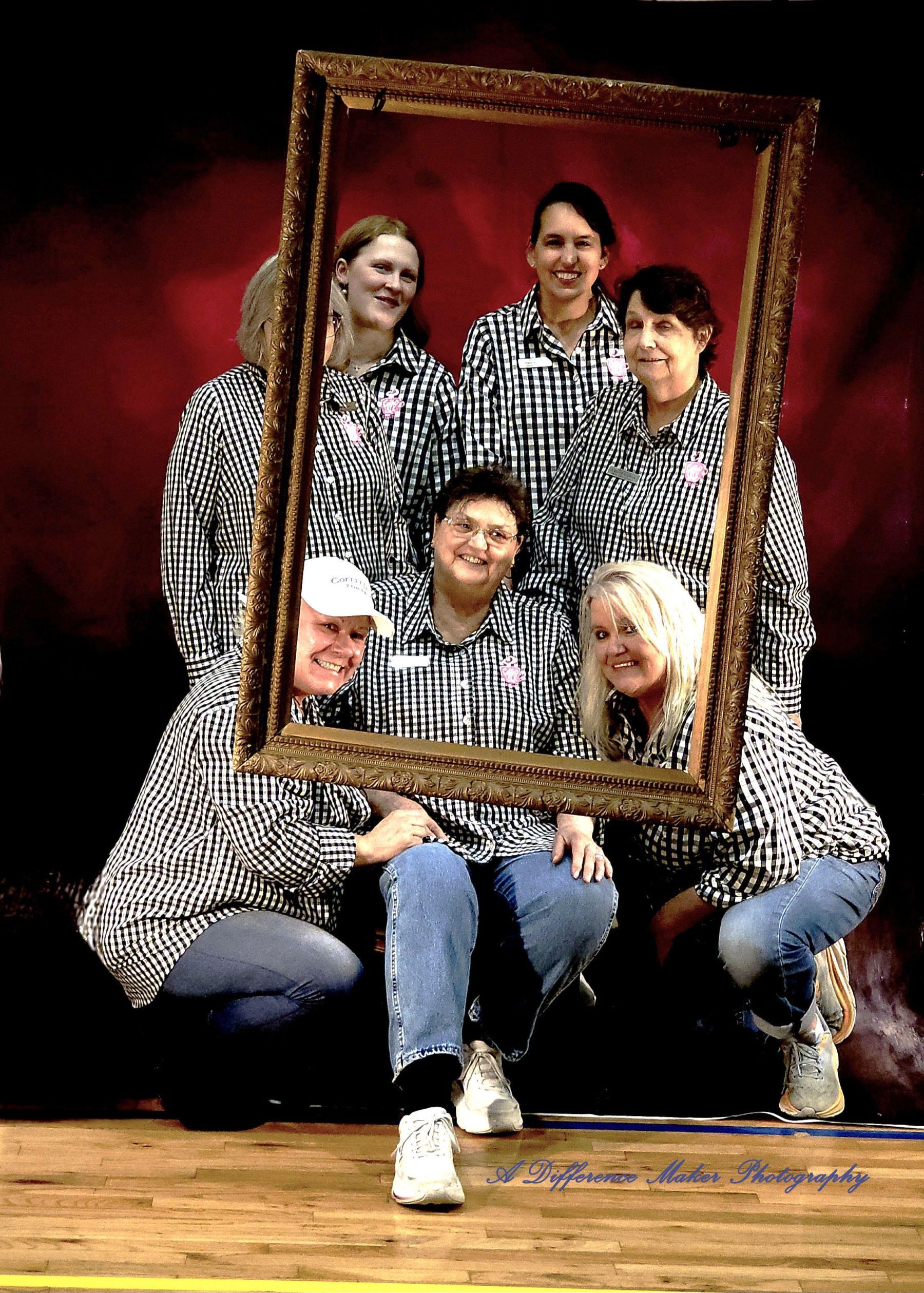 Group of people in matching shirts posing with a frame. They're on a stage with a red backdrop.