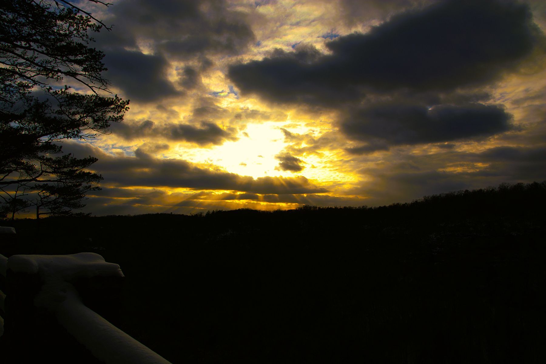 Sunset through clouds over a dark landscape, with a hand holding a rail in the foreground.