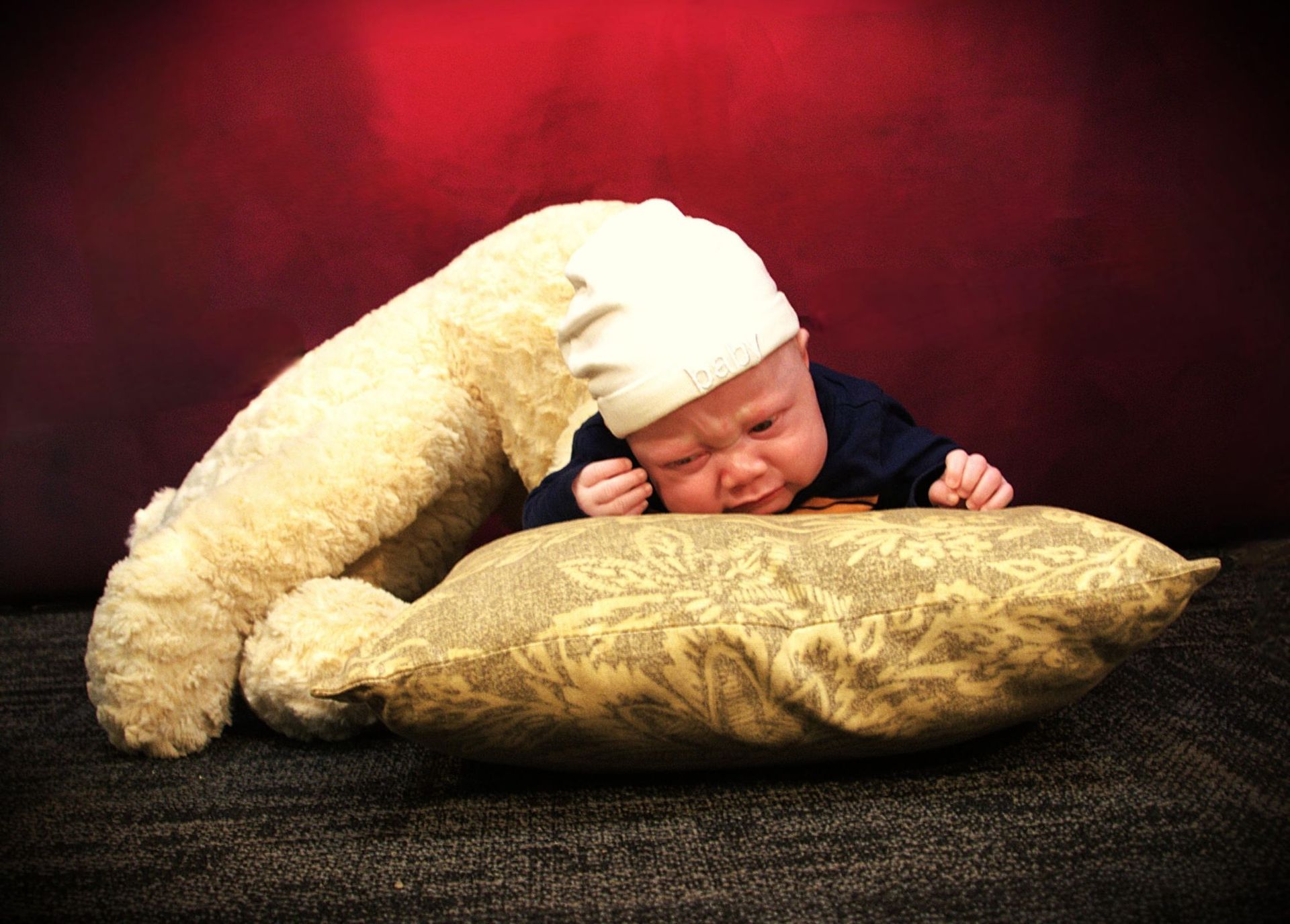 A baby wearing a white beanie is laying on a patterned pillow next to a stuffed bear, face scrunched with a frown.