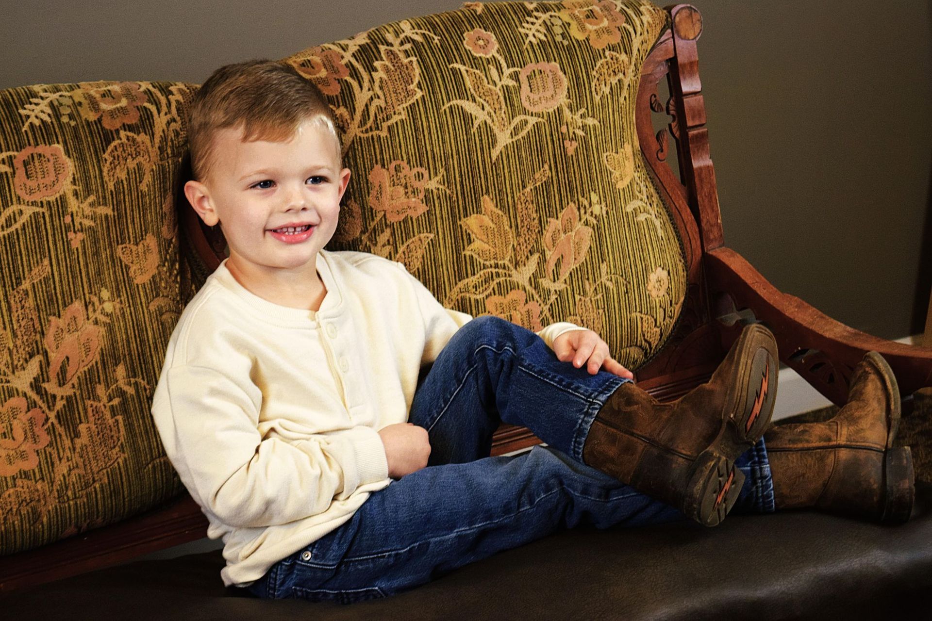Boy in jeans and cowboy boots smiles, sitting on a patterned couch.