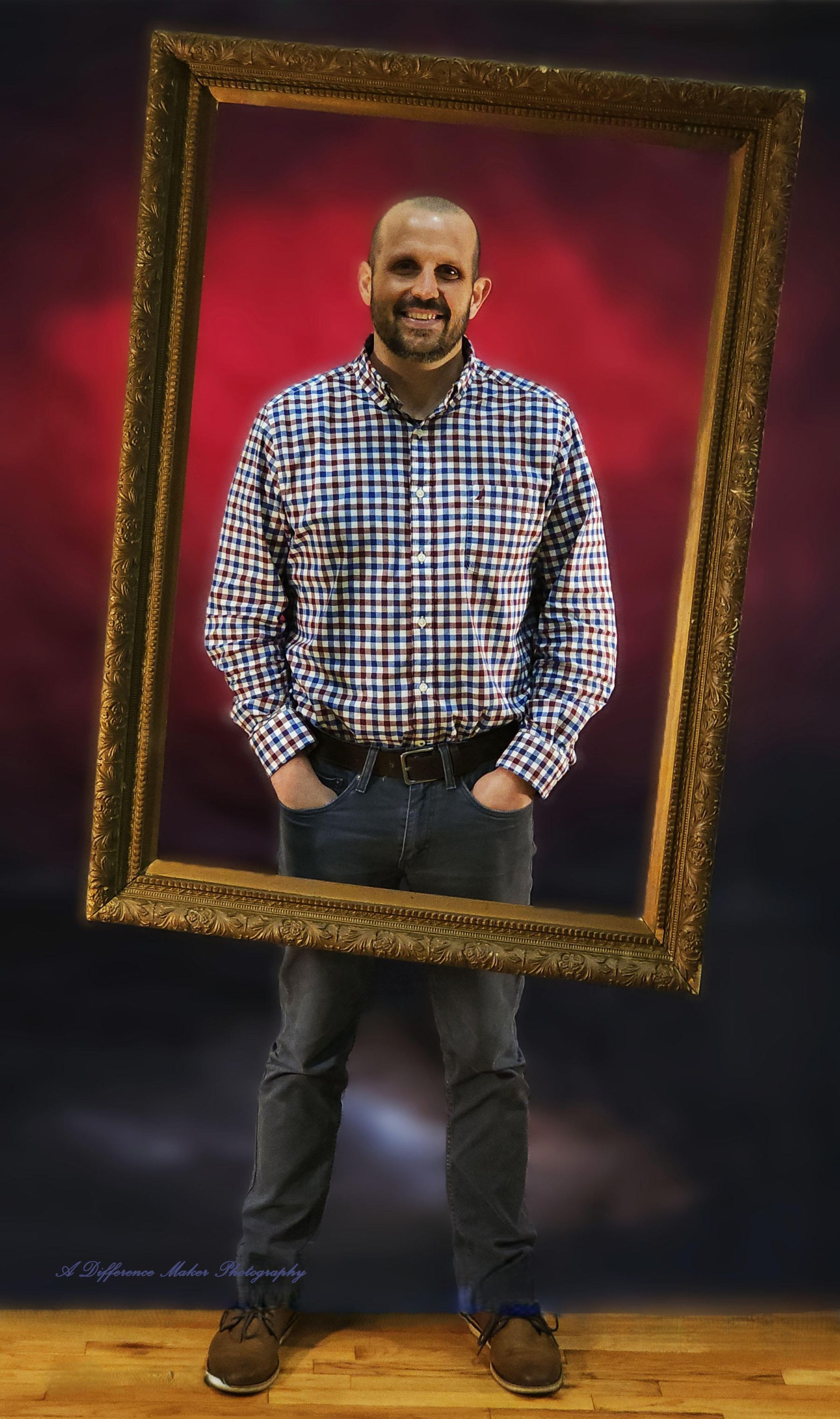 Man framed by ornate gold frame, standing, hands in pockets. Plaid shirt, jeans, red backdrop.