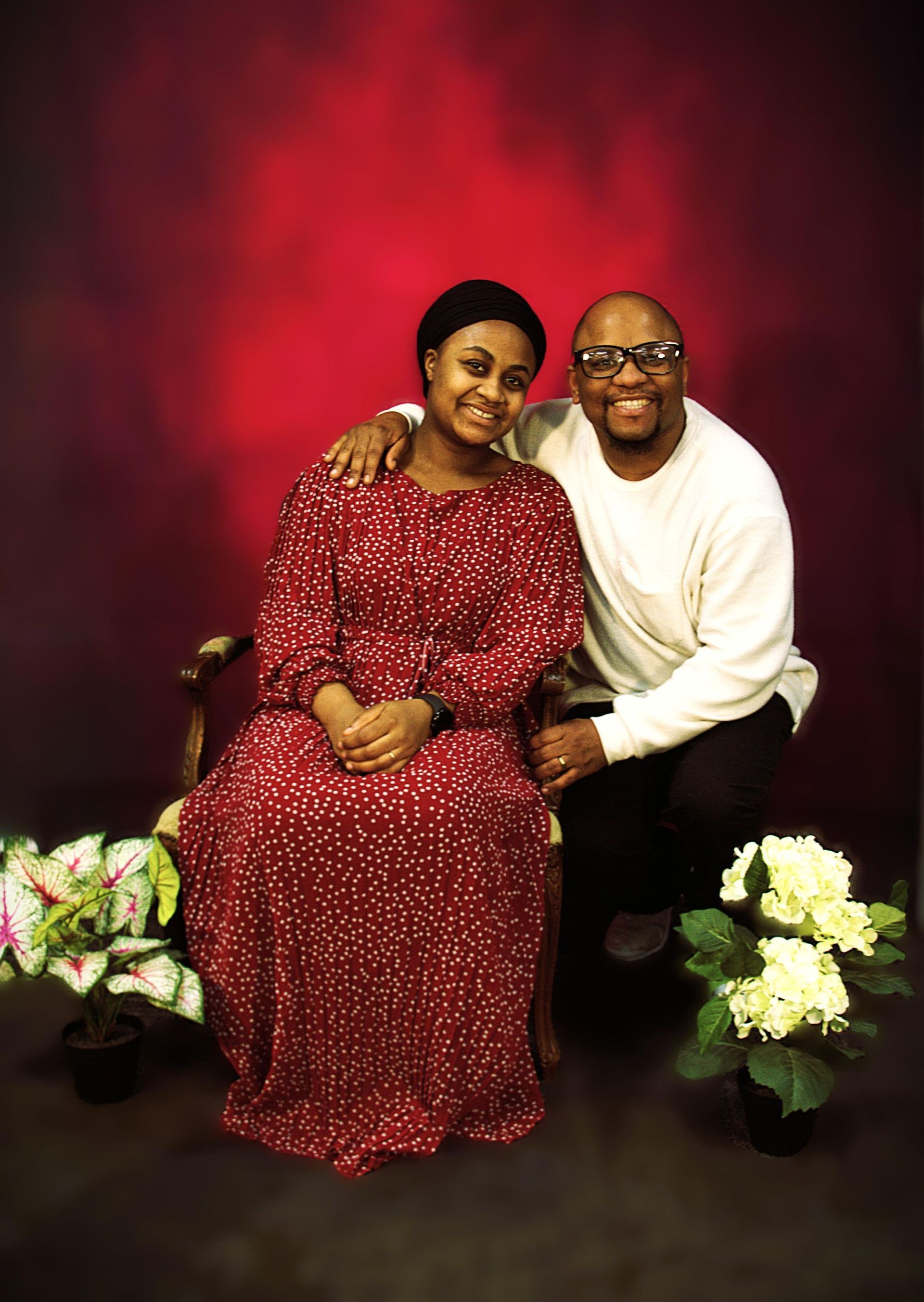 Couple poses smiling. Woman in red dress sits, man in white sweater stands, arm around her. Red background.