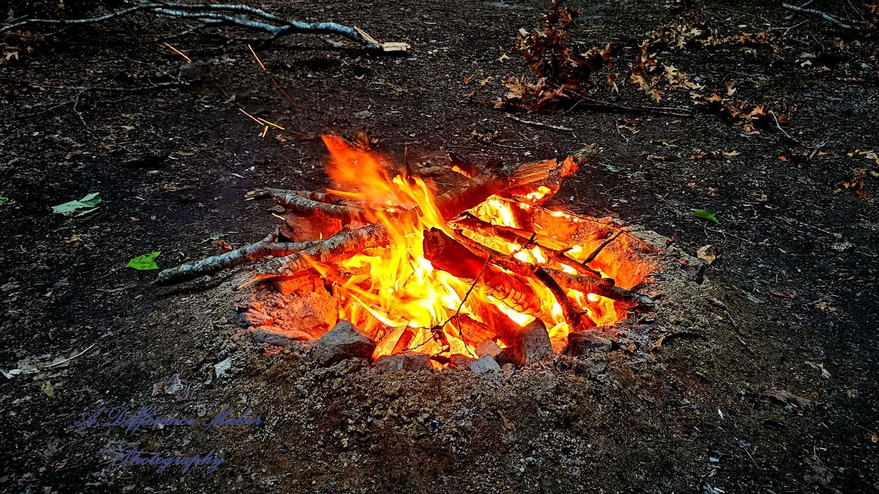 Campfire burning with orange and yellow flames, surrounded by dark ground and wood.