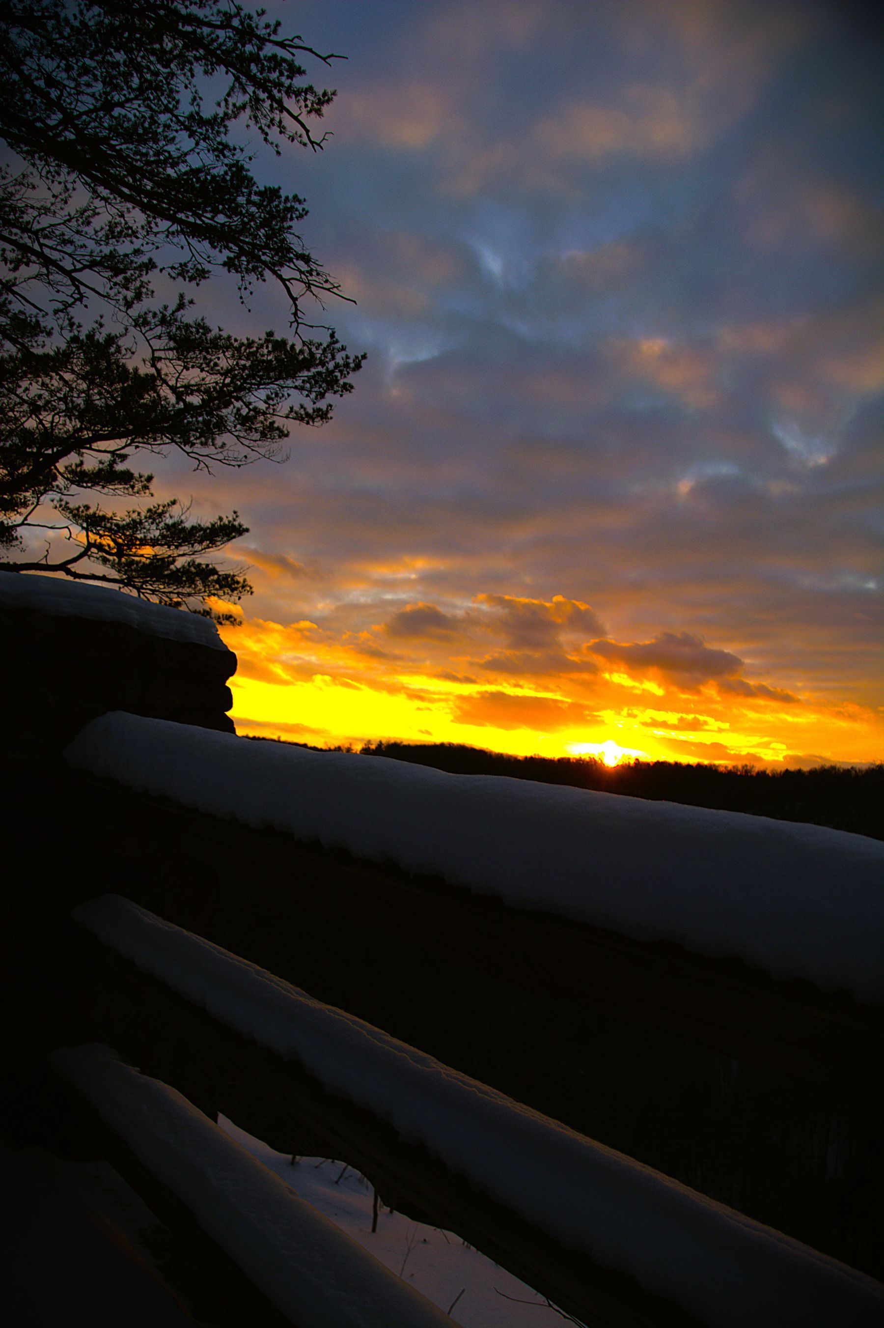 Sunset over snowy landscape, vibrant orange and yellow hues in sky, dark silhouetted trees and fence.