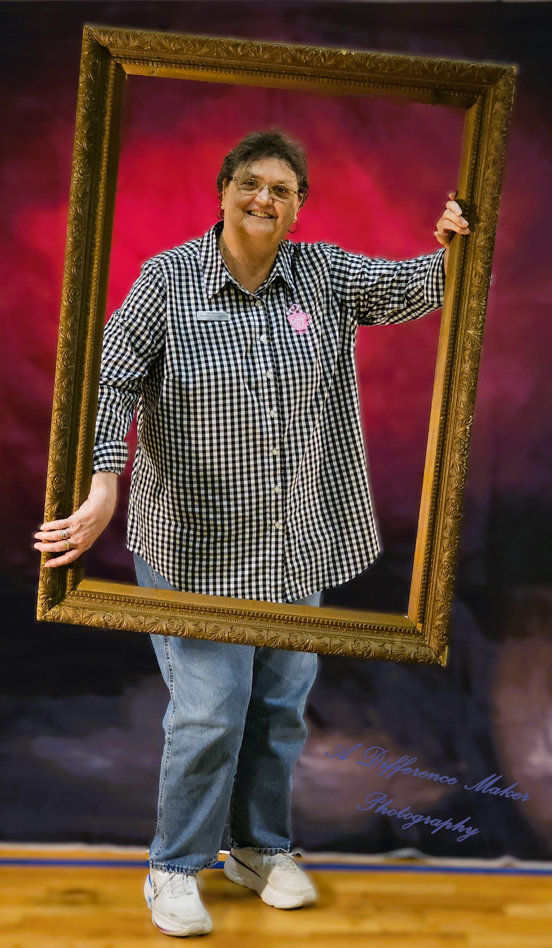 Woman holding a large, ornate golden frame, smiling. Wearing a checkered shirt and jeans, standing against a dark background.
