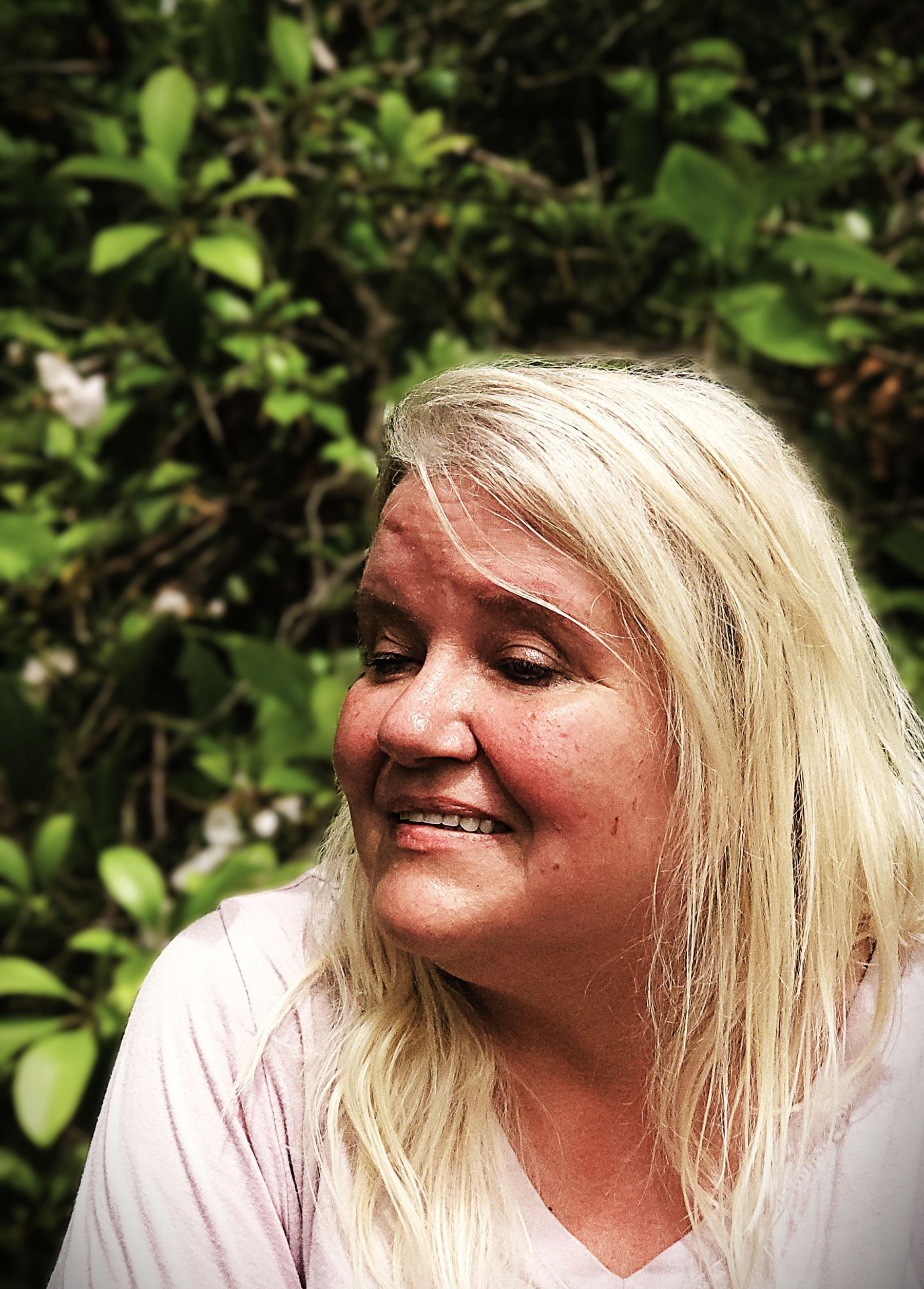 Blond-haired person smiles, looking to the side, against a blurred green leafy background.