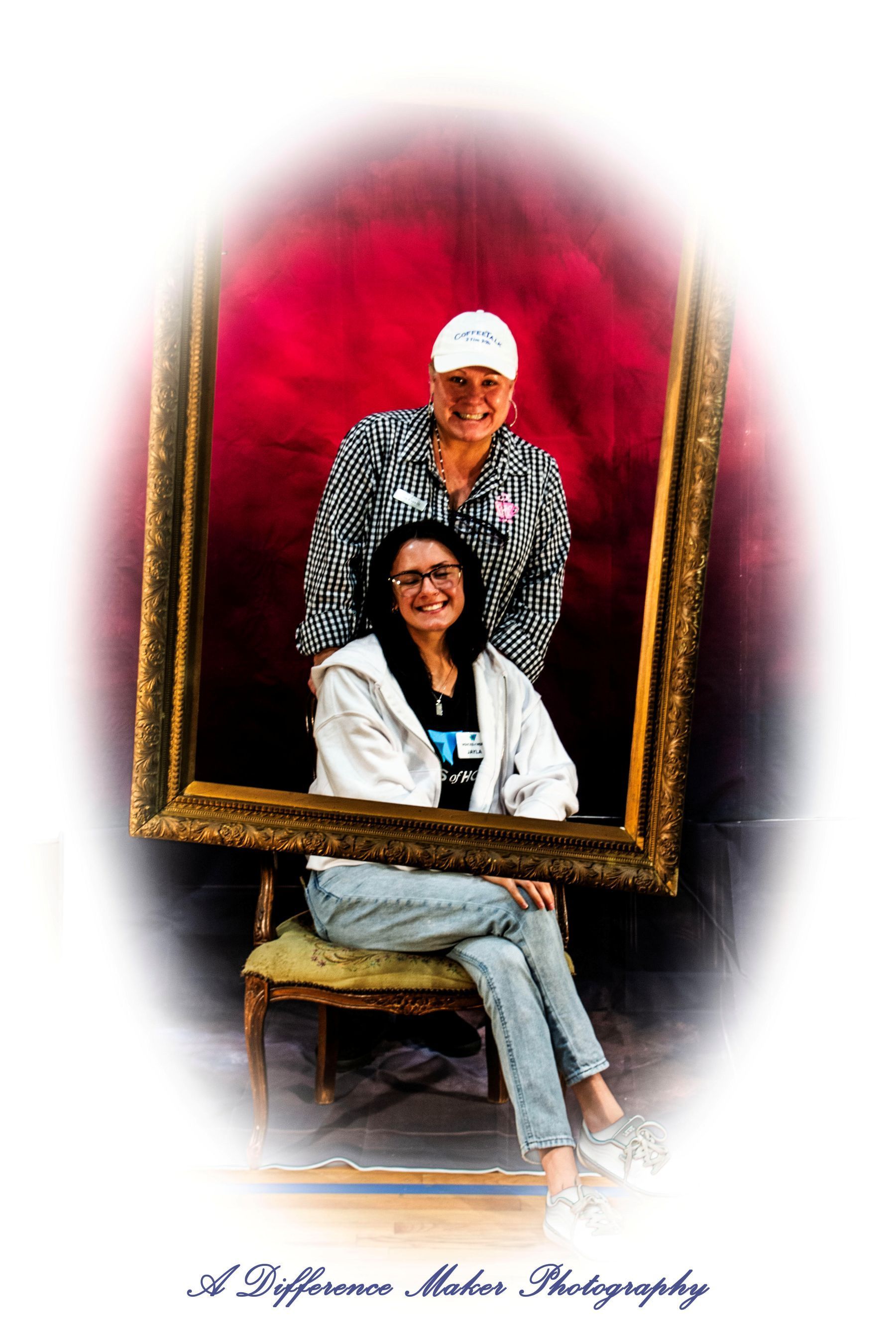 Two people posing inside a gold picture frame. One is seated, the other stands behind wearing a white hat, red backdrop.