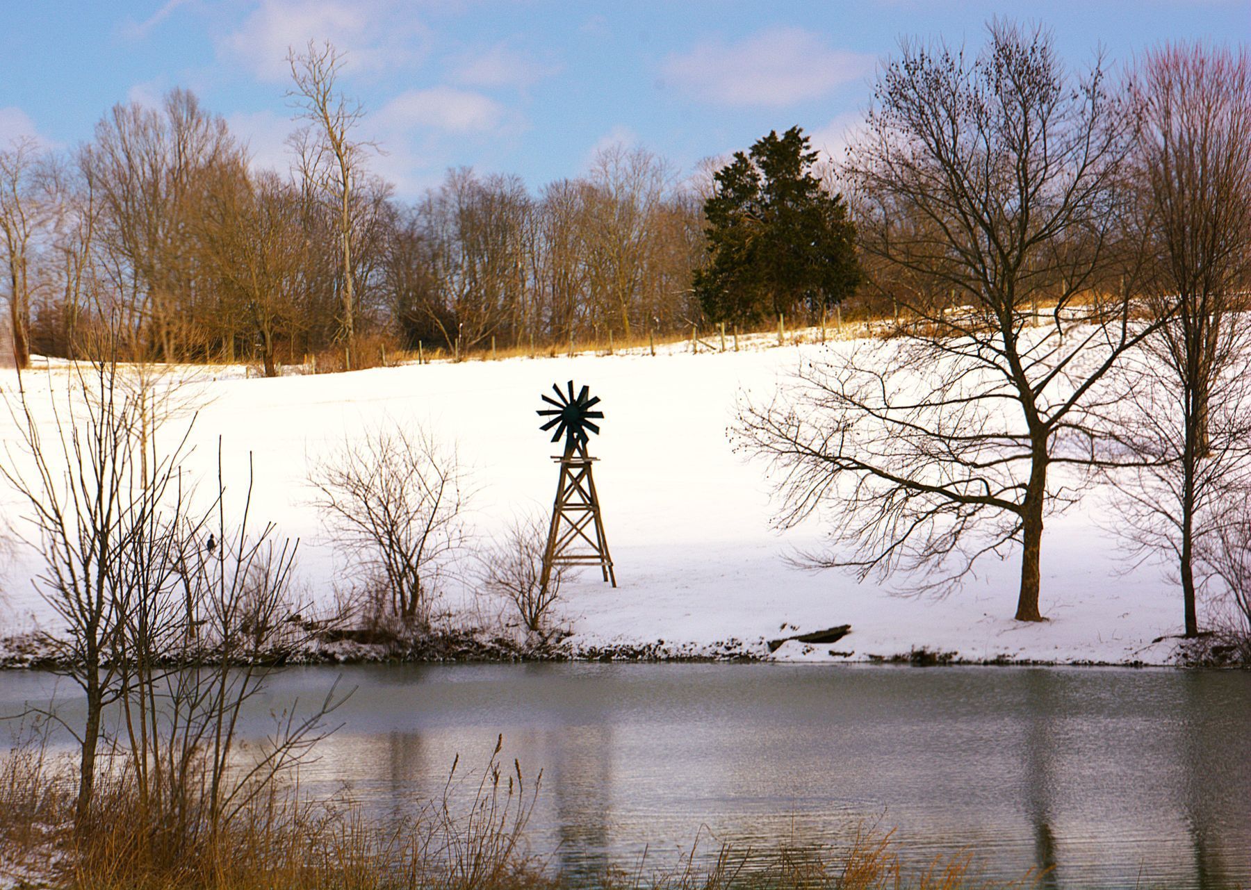 Snow-covered hillside with pond, windmill, and leafless trees under a blue sky.