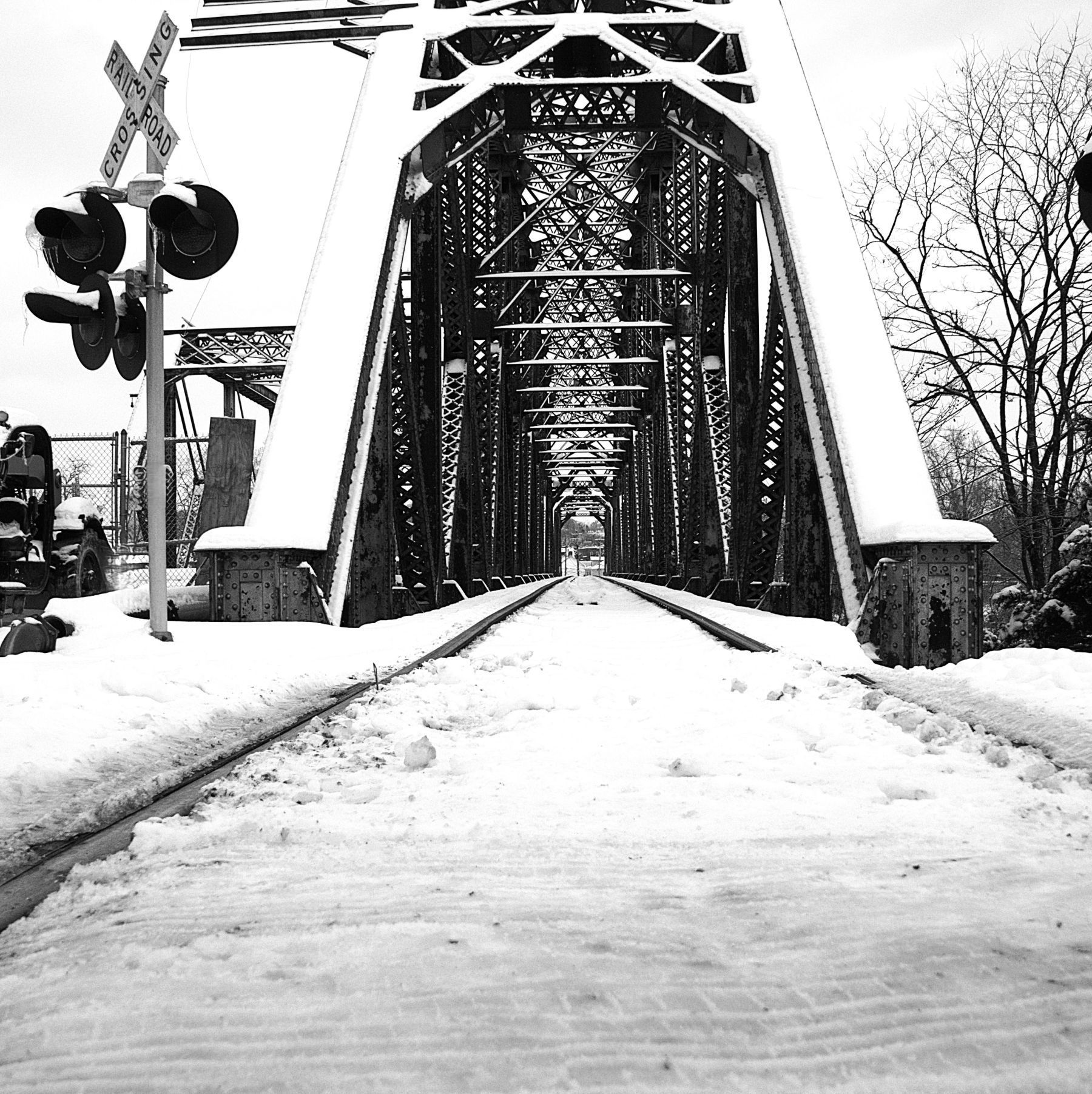 Snow-covered train tracks through a steel truss bridge, crossing sign and signal in view. Black and white.