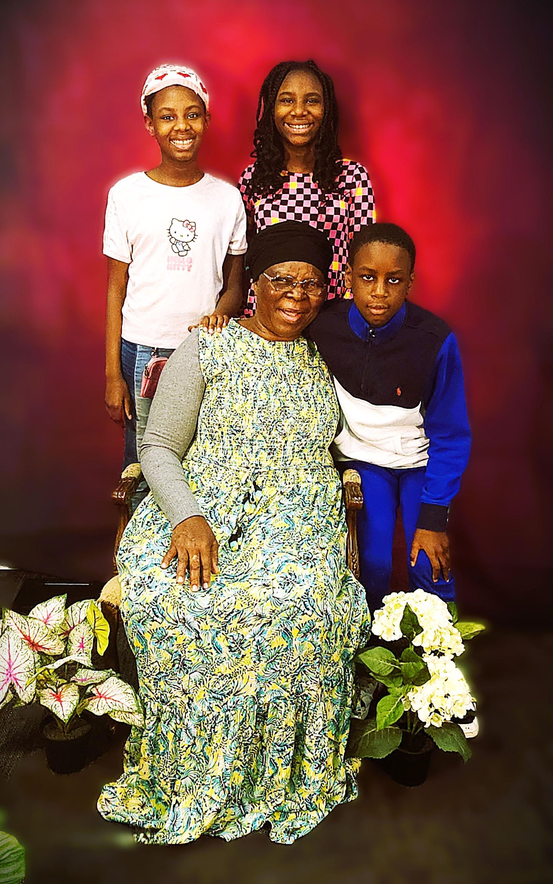Woman seated with three others, all smiling, in front of a red backdrop, near floral arrangements.