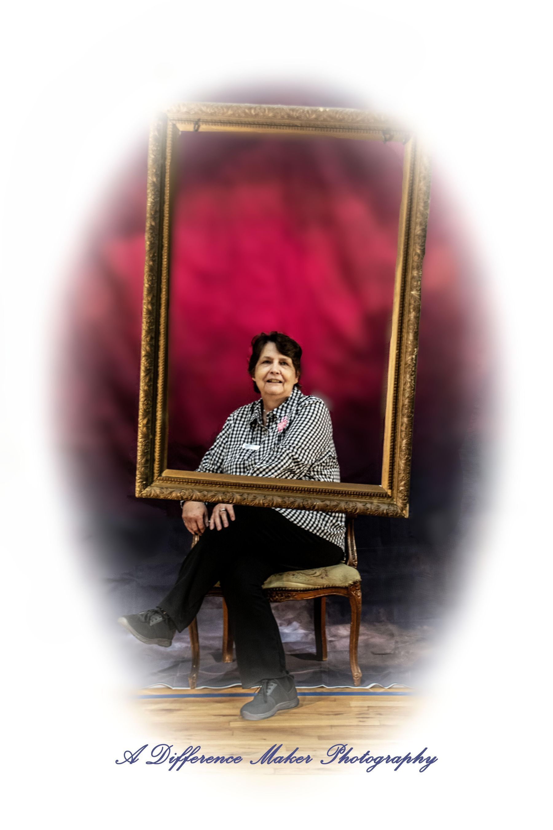 Woman seated, framed by a large gold ornate border against a red backdrop. Wearing black and white top and black pants.