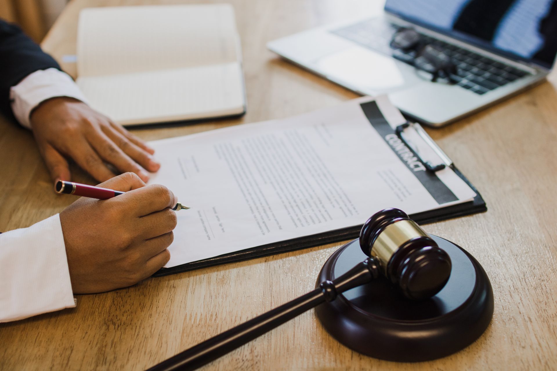 Person signing a legal contract beside a judge’s gavel on a wooden desk. Person signing a legal contract beside a judge’s gavel on a wooden desk.