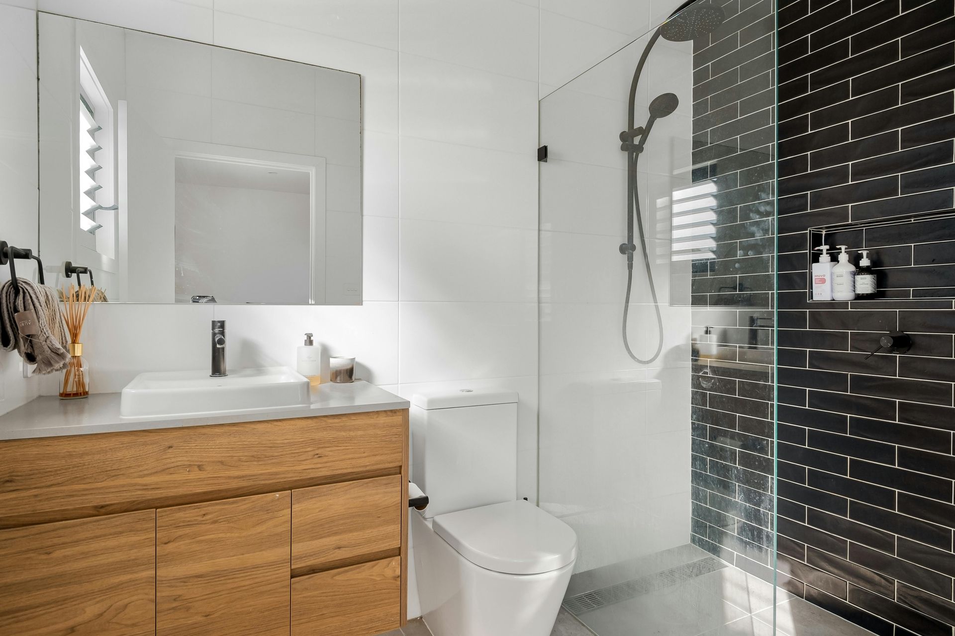 Modern bathroom with wood vanity, white walls, black tile shower, glass door, and toilet.