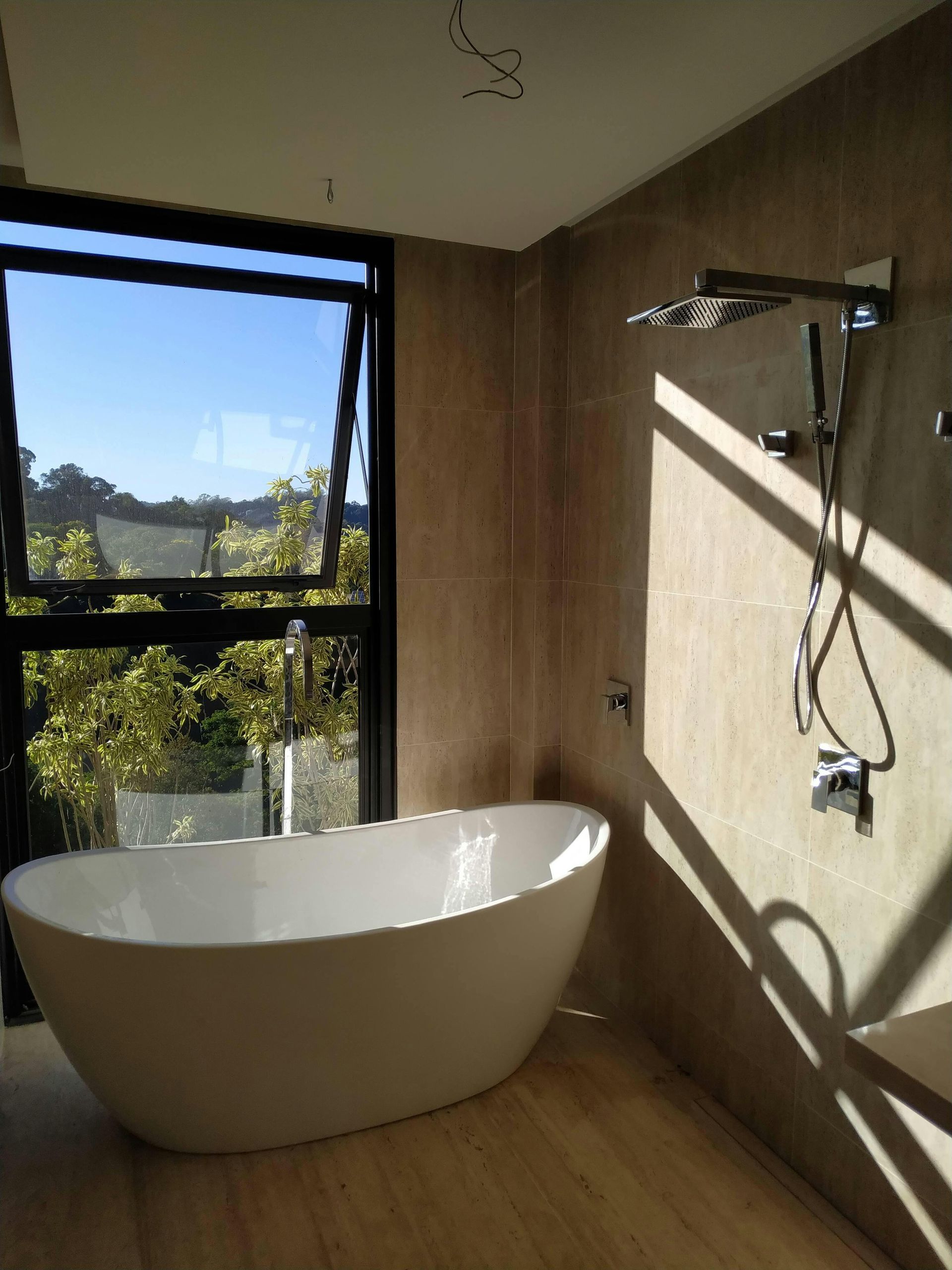 Bathroom with a white freestanding tub, large window, and shower fixtures on a textured wall; sunlight streams in.