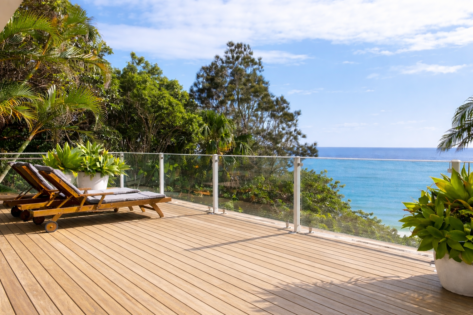 Wooden deck with ocean view, two lounge chairs, glass railing, sunny sky, lush greenery.