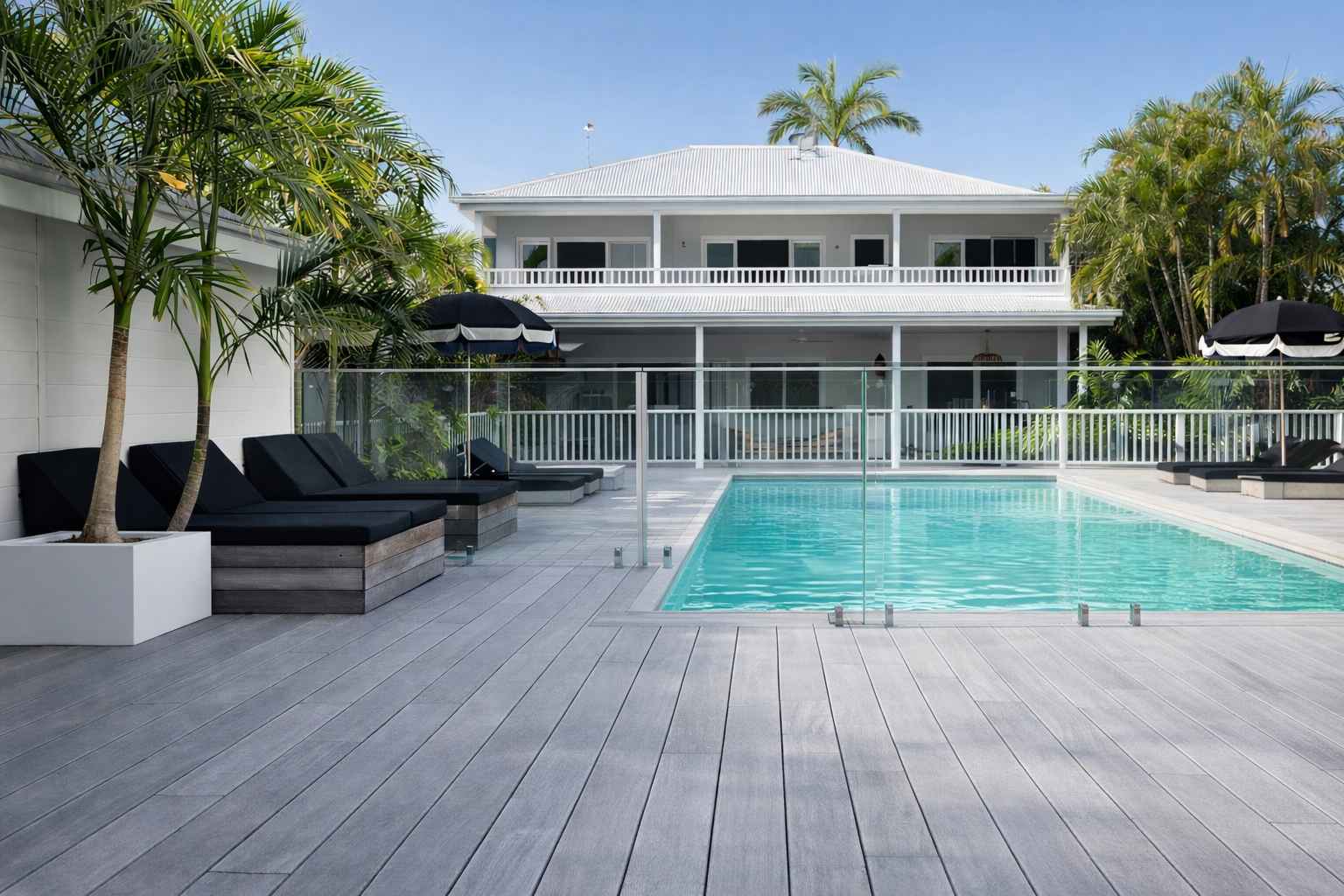 Two-story white house with pool and deck; black lounge chairs and umbrellas.