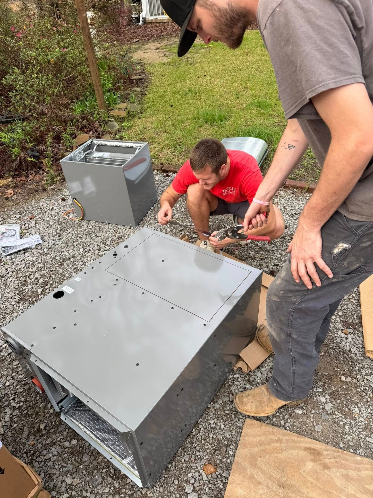 Two people work together to assemble a large, gray metal HVAC unit outdoors on a gravel surface.