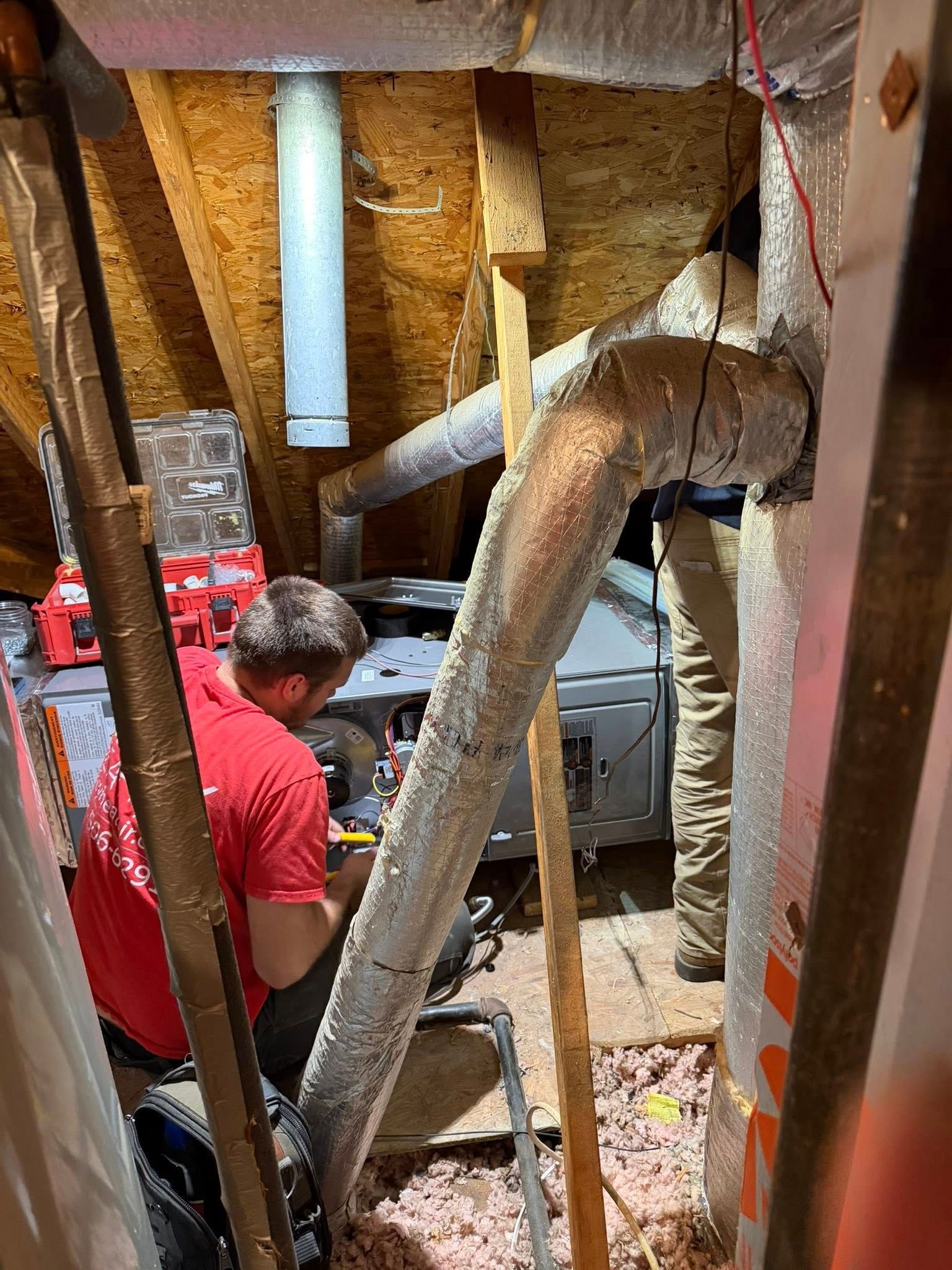 Two technicians in an attic perform maintenance on a furnace unit, surrounded by silver ducting and insulation material.