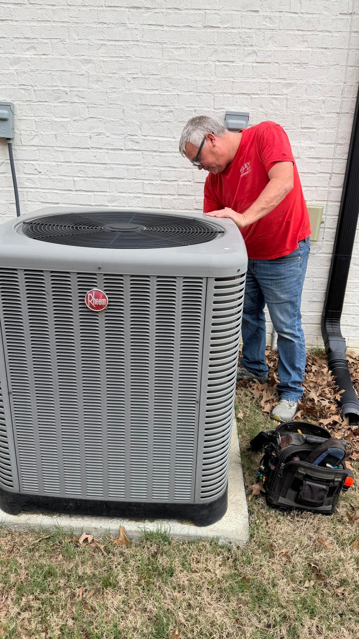 A person in a red shirt works on an outdoor HVAC unit next to a light-colored brick wall.