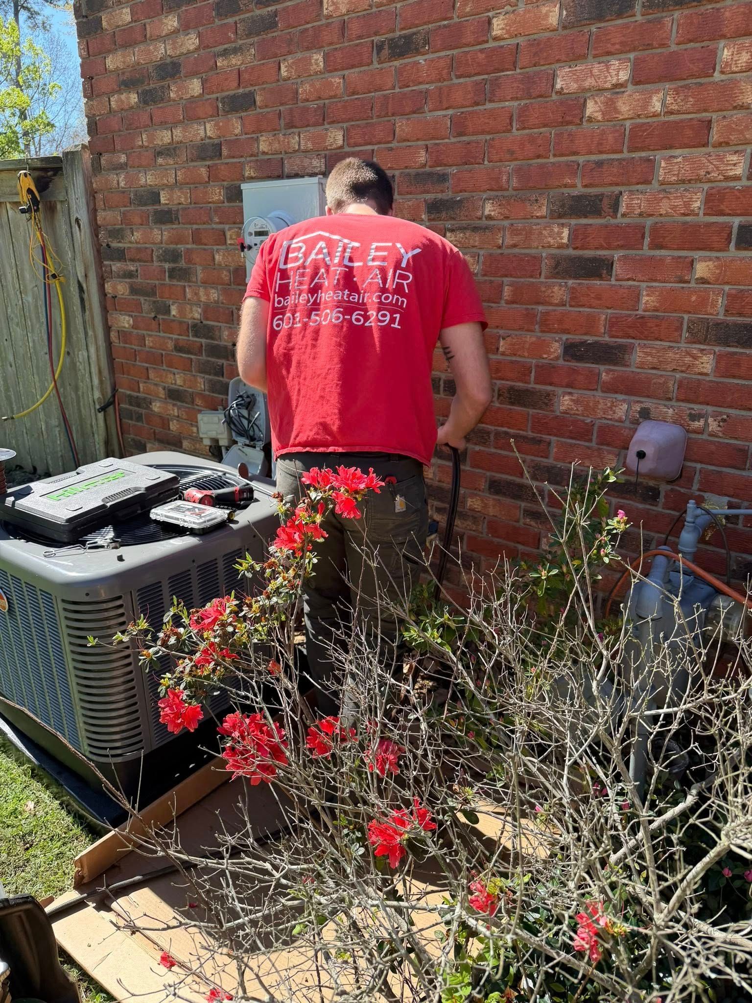 A technician in a red t-shirt works on an outdoor air conditioning unit next to a brick wall.