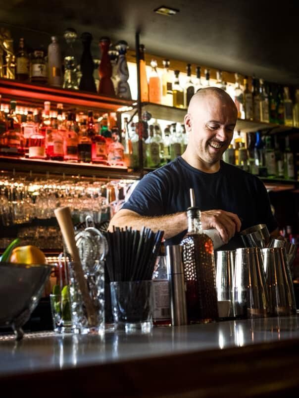 Bartender smiles, mixing drinks behind a bar with liquor bottles, glasses, and a muddler.