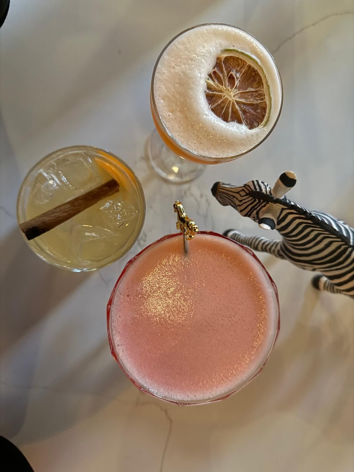Three cocktails on a white surface, one with a dried orange slice, zebra figurine.