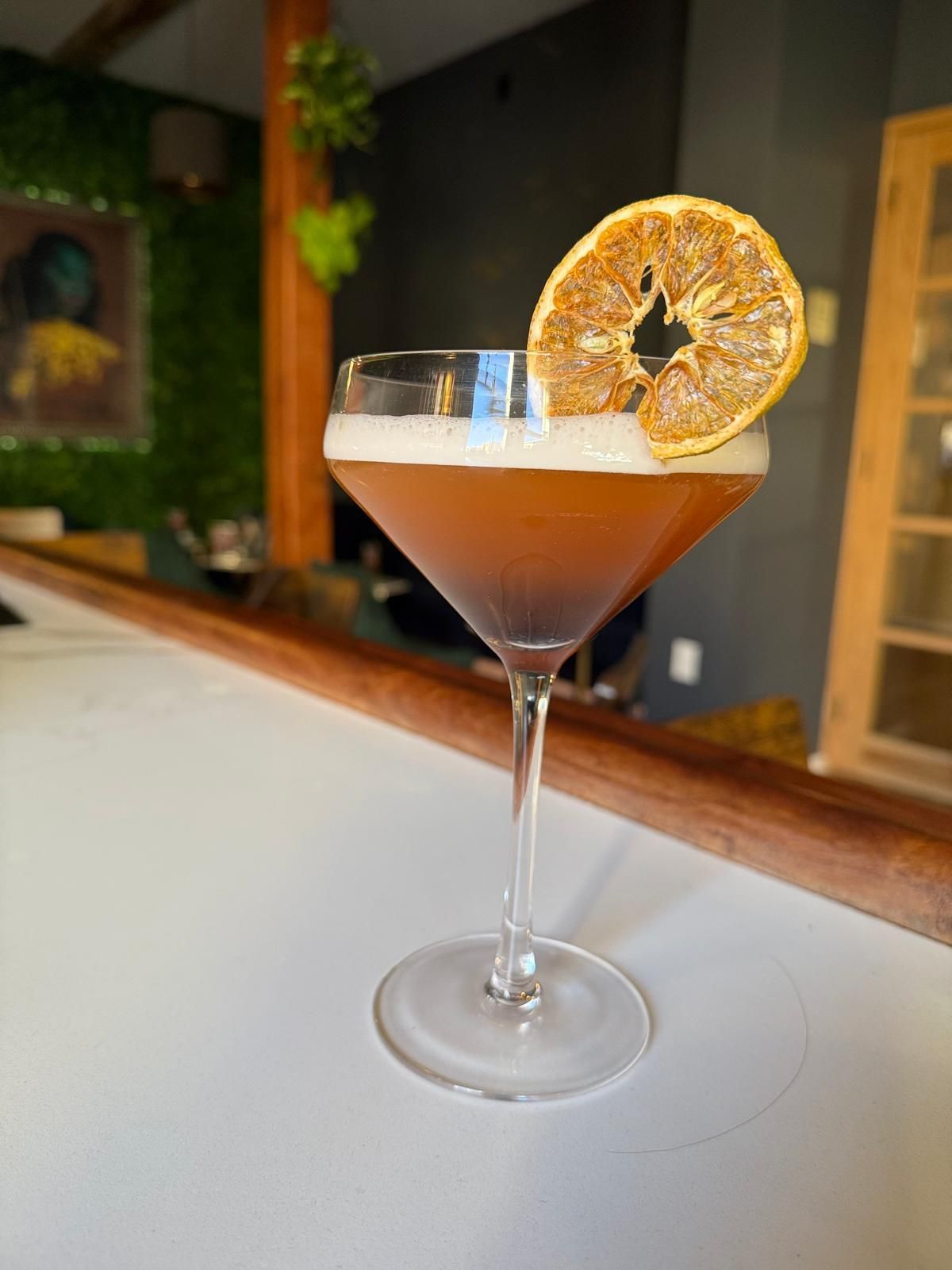 Cocktail in a stemmed glass, garnished with dried orange slice, on a white bar.
