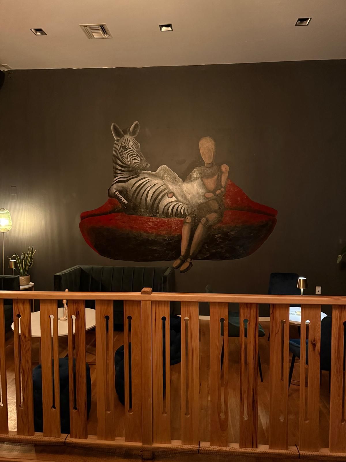 Mural of a zebra and person lounging on a red lip against a dark wall. Wood railing in the foreground.