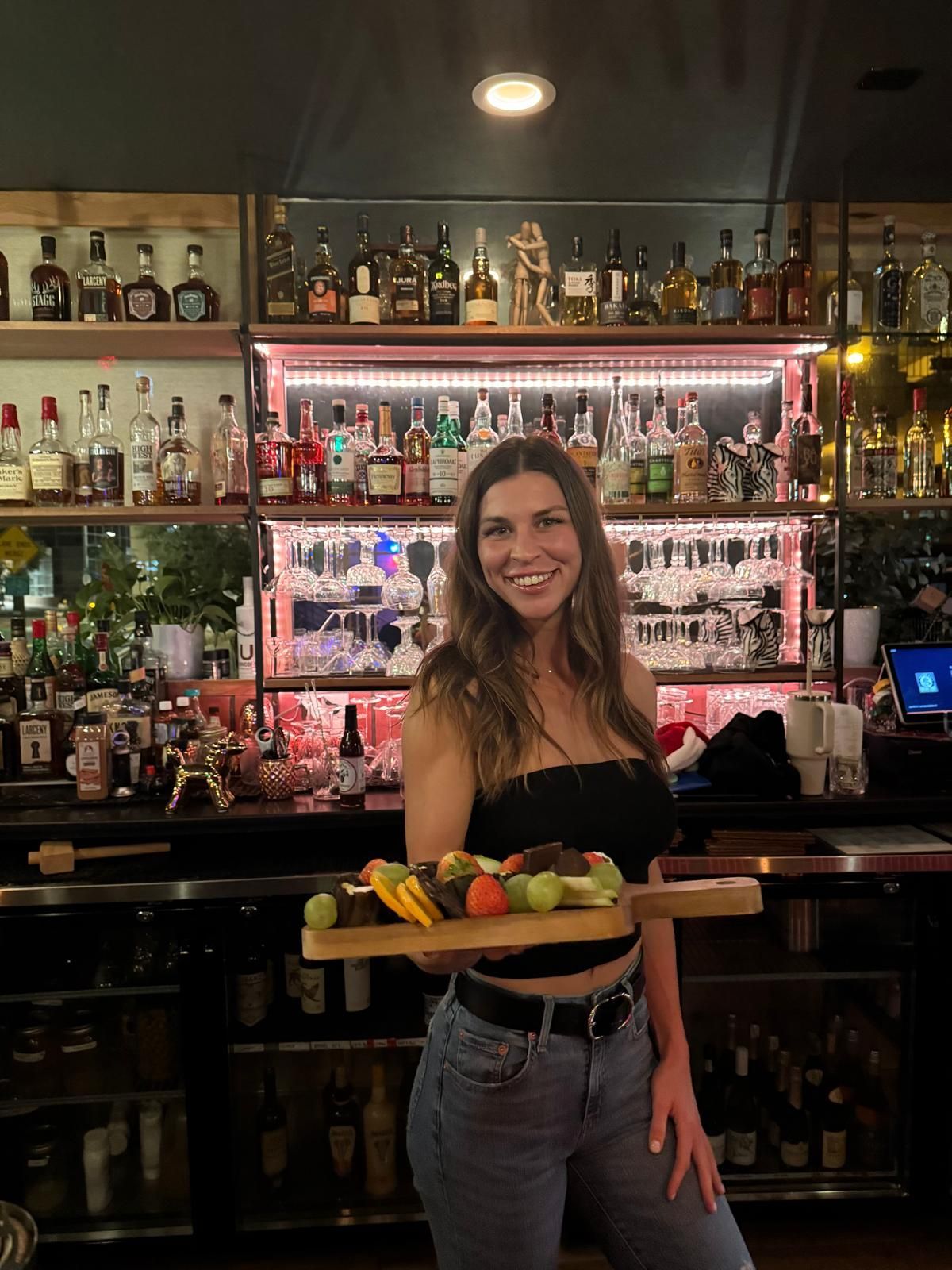 Woman holding a wooden board with food, smiling at the camera in front of a bar.