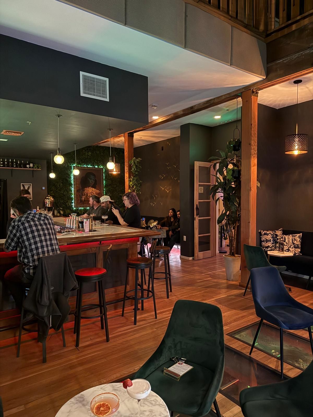 Interior of a bar with people at the bar and seating areas, wooden floors, and dark walls.