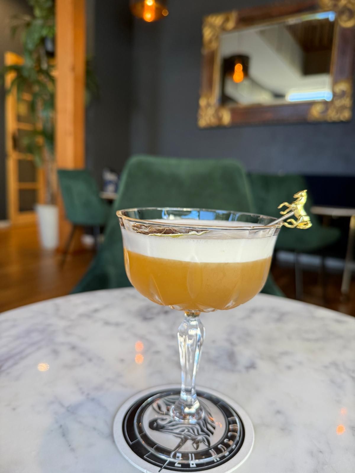 Cocktail in a stemmed glass with foamy top, on a marble table with a coaster in a dimly lit bar.