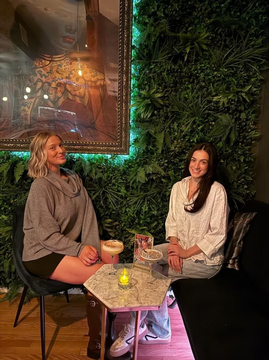 Two women seated at a table, smiling. One in gray sweater, the other in white, with a green wall backdrop.