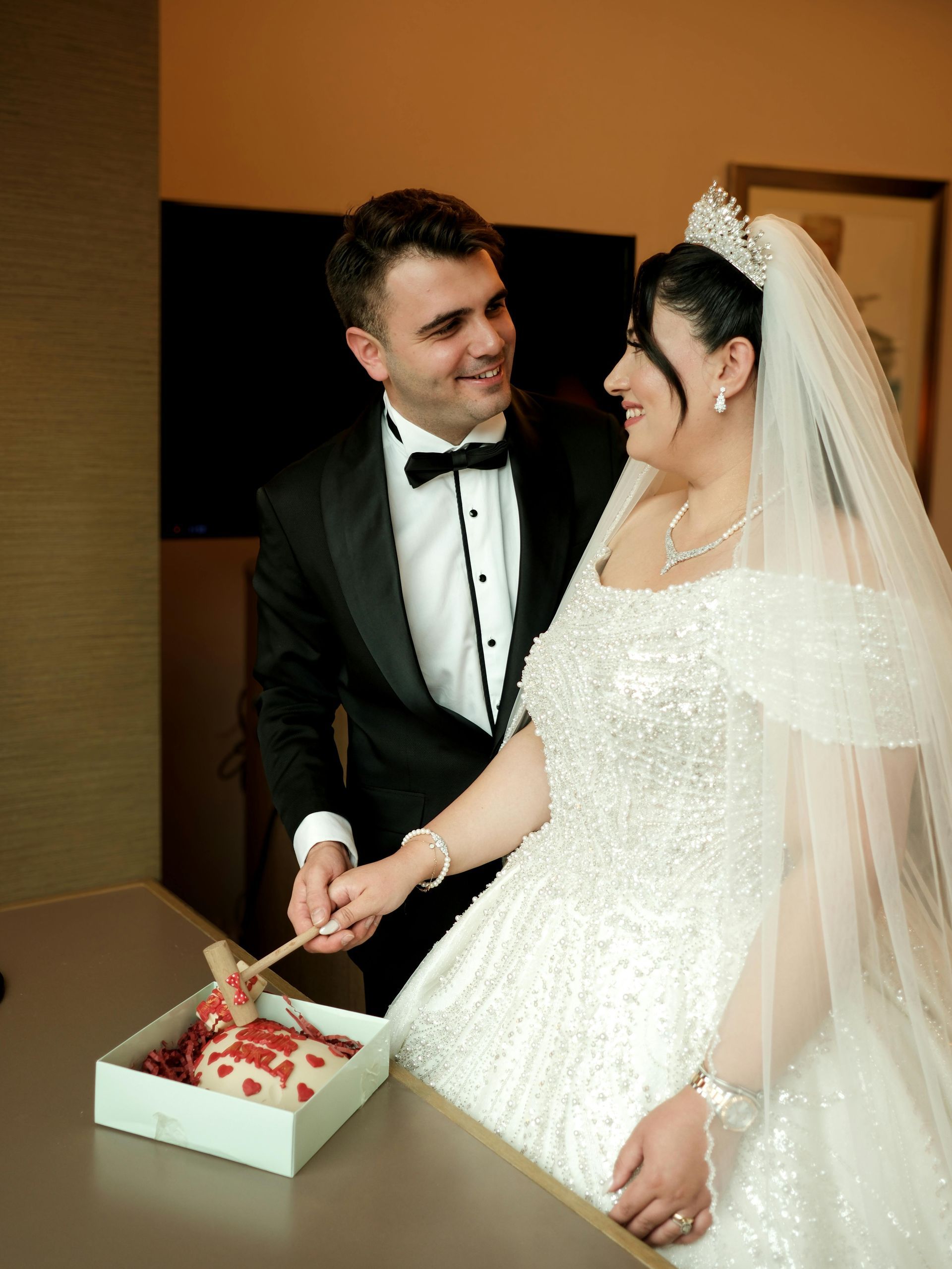 Bride and groom cutting wedding cake. Man in tuxedo, woman in beaded gown with veil.