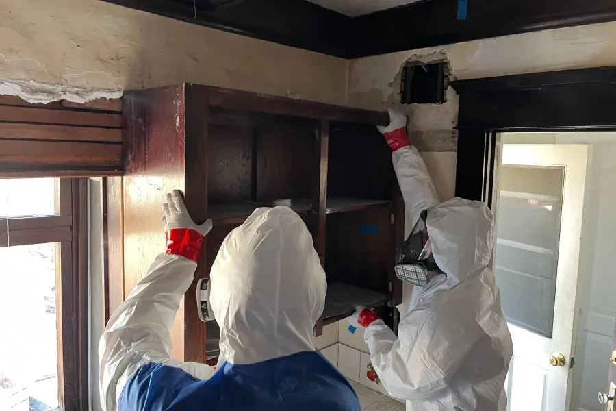Two people in protective suits are working on a shelf in a room.