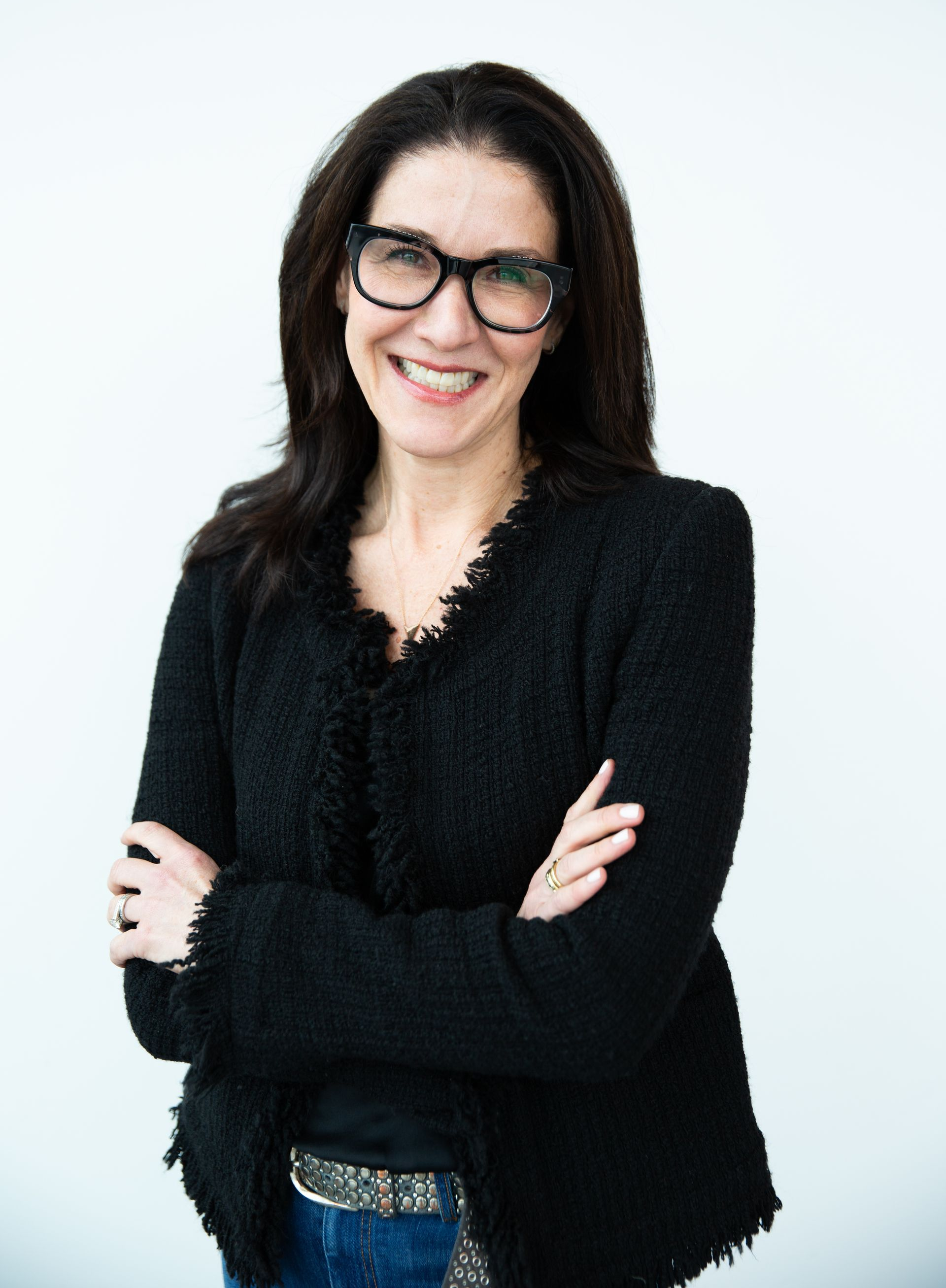 Woman with black hair, glasses, and jacket, arms crossed, smiles.